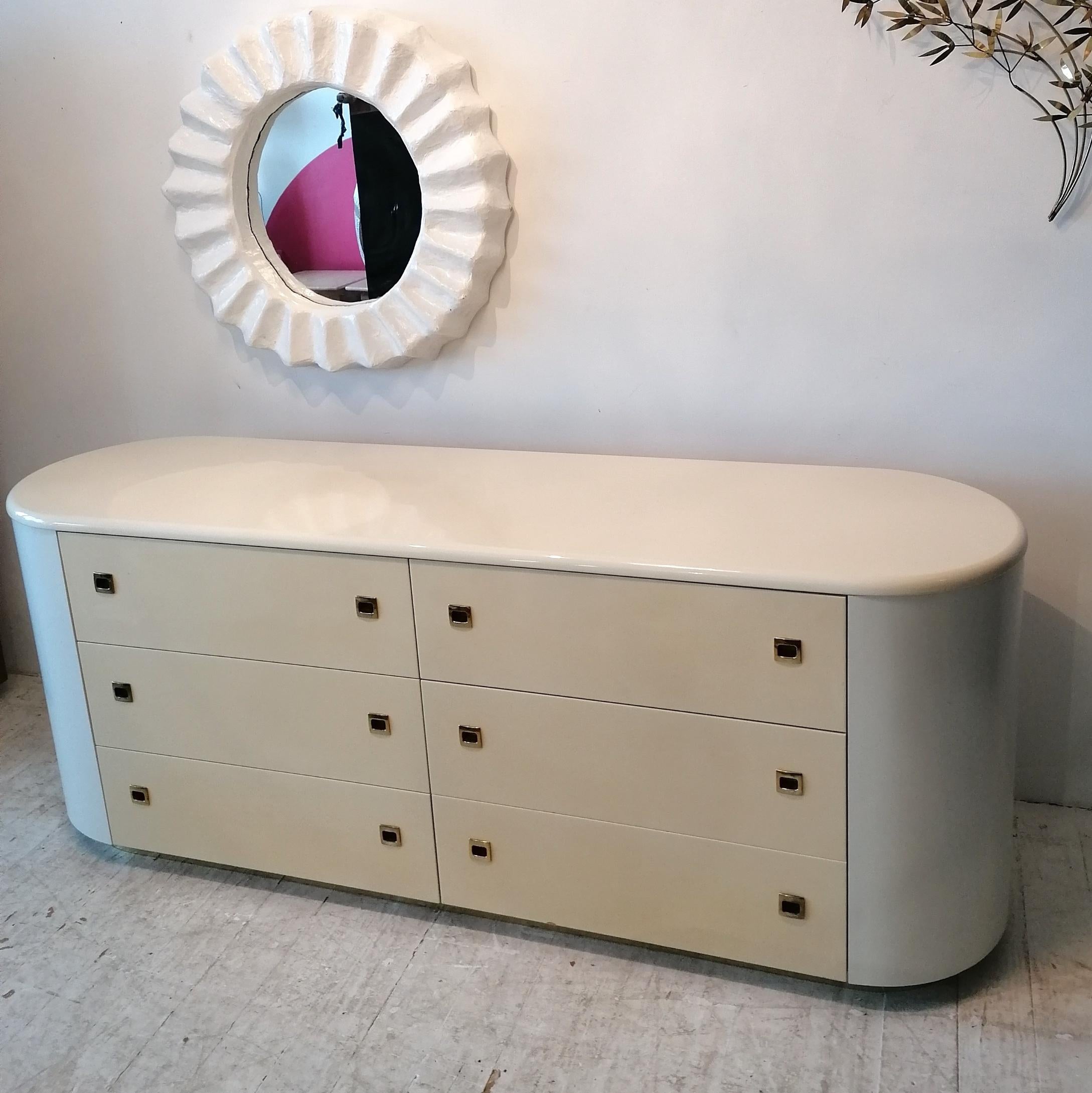 Gorgeous pill-shaped Deco Revival sideboard or dresser with six large drawers, 1980s American. 
Drawer fronts are shiny cream lacquer, the rest of the piece is off-white, with a soft sheen. Drawer handles are gold metal and the plinth is also gold