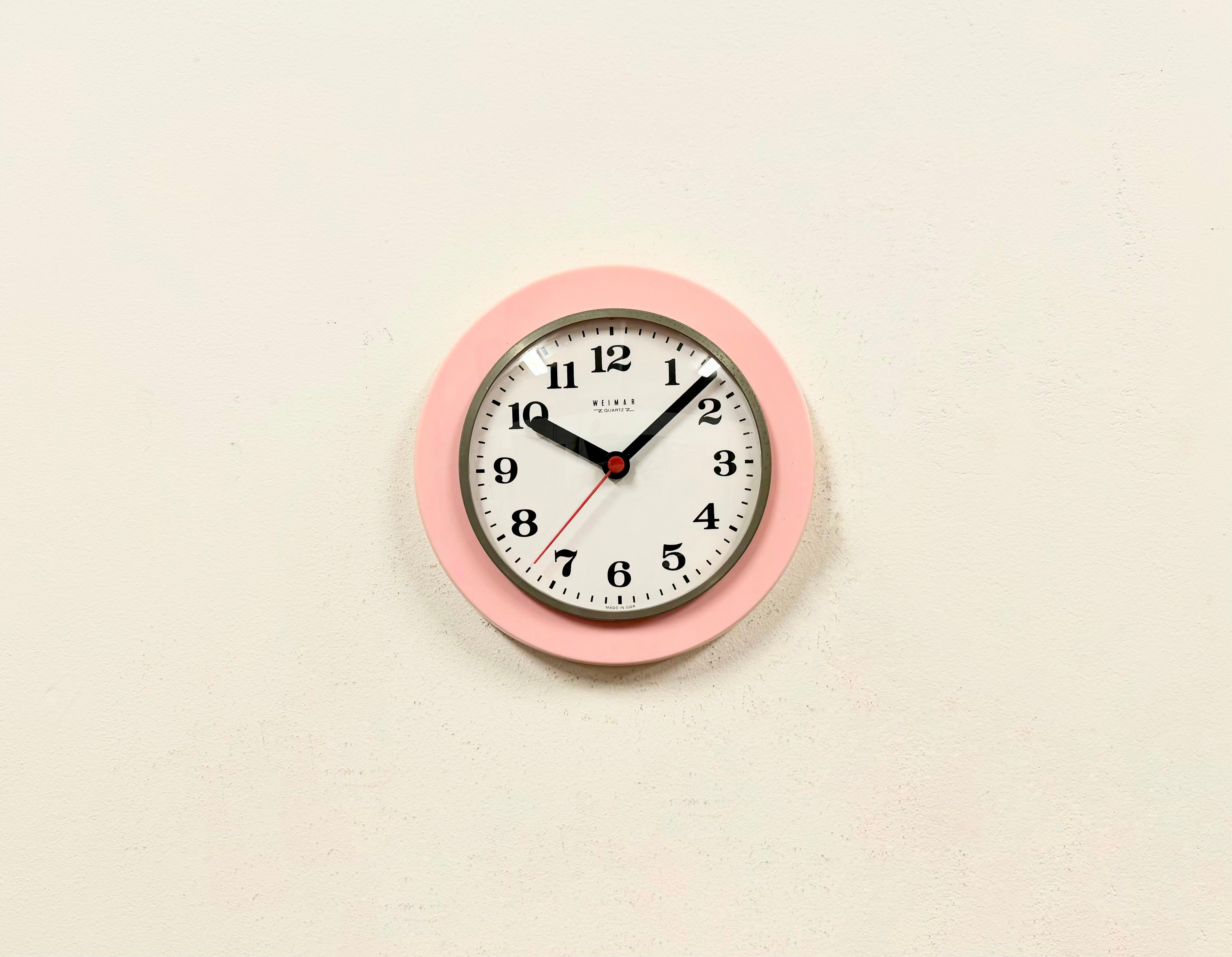 Vintage industrial wall clock produced by Weimar Quartz in former East Germany during the 1980s. It features a pink bakelite body, an aluminium hands and a curved clear glass cover with metal ring. Battery-powered clockwork requires 1 AA battery.