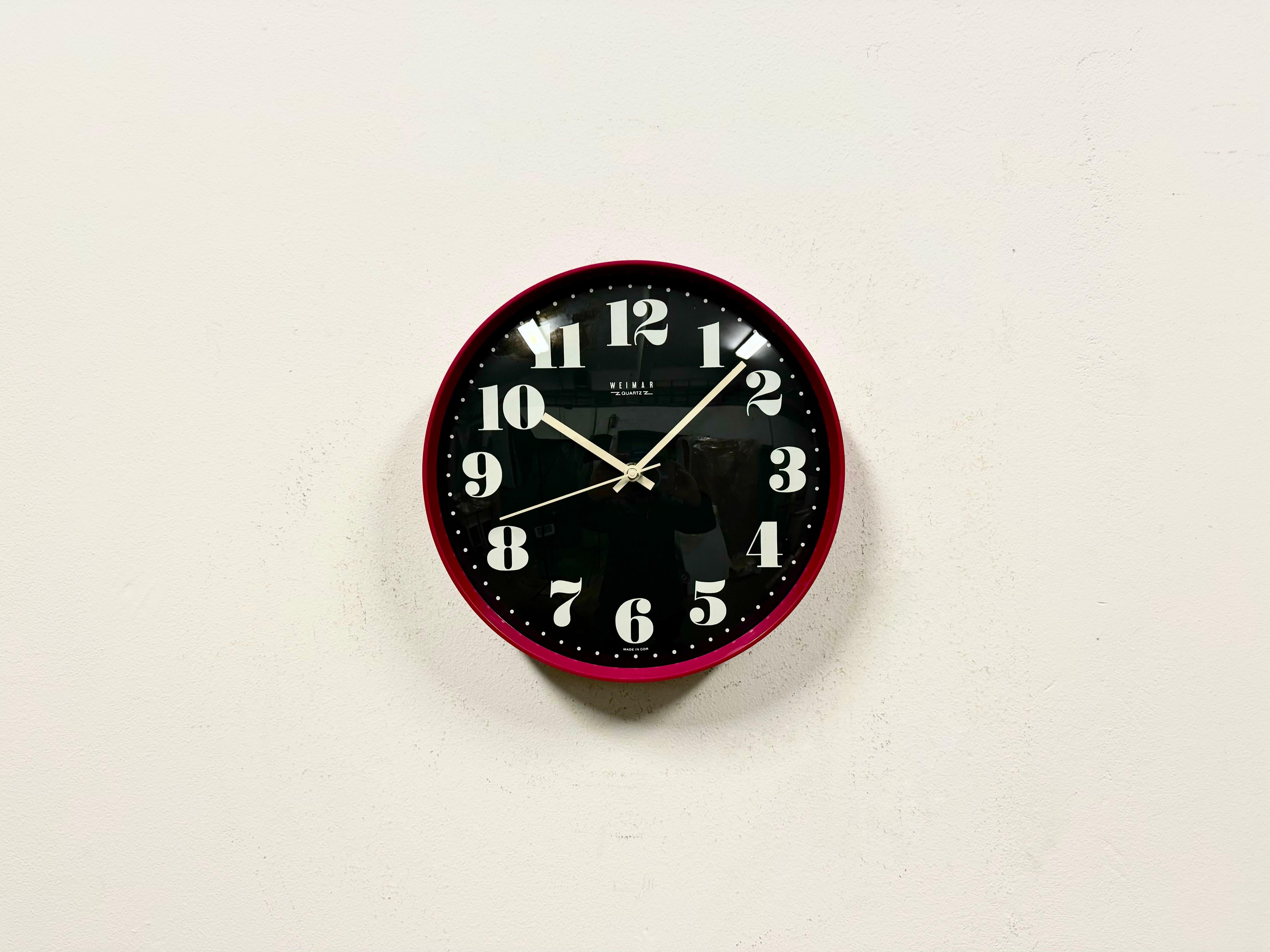 Vintage industrial wall clock produced by Weimar Quartz in former East Germany during the 1980s. It features a pink bakelite frame,a black metal dial, an aluminium hands and a curved clear glass cover. The original battery-powered clockwork requires