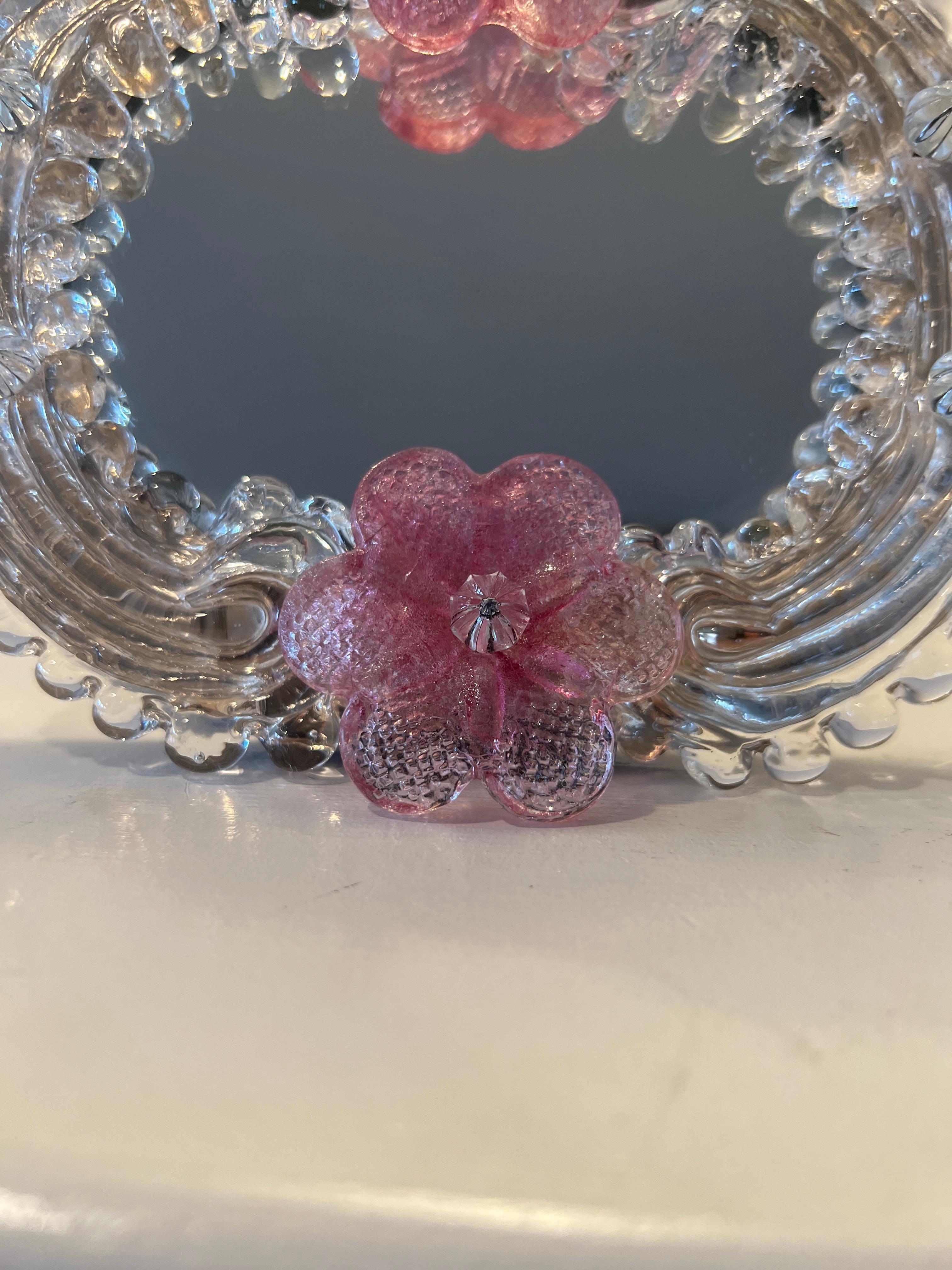 Venetian, mid 20th century.

A petite art glass mirror made with two pink Venetian glass florets mounted to around a gold freckled scalloped edge frame. Verso has a stand for using on table top and a hook for wall use. Unmarked.  