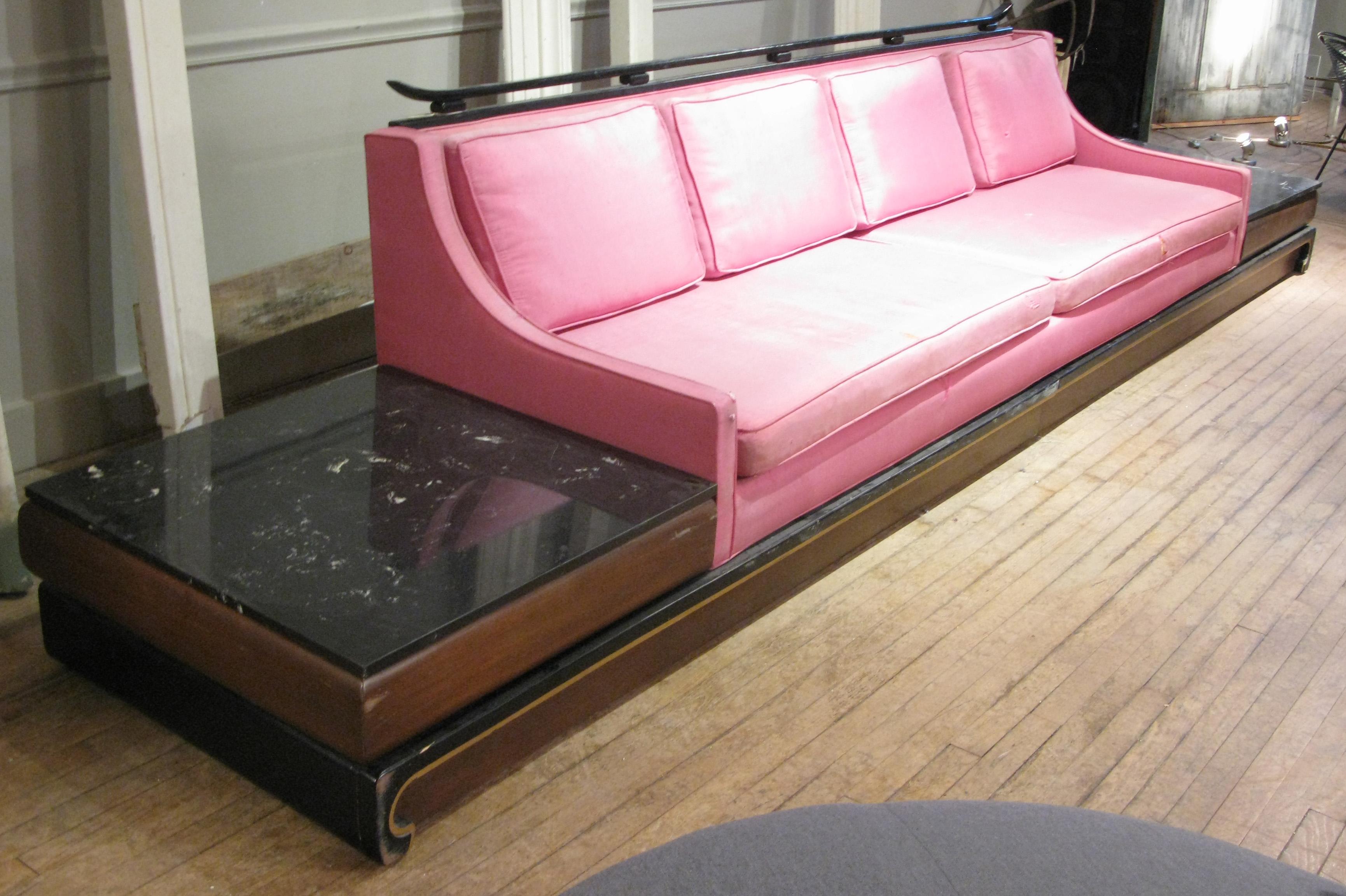 A vintage 1960s long platform base sofa with integrated marble-top tables at either end. The base and backrest have Asian inspired details. The sofa covered in its original pink silk, which will need to be redone. The frame shows age expected