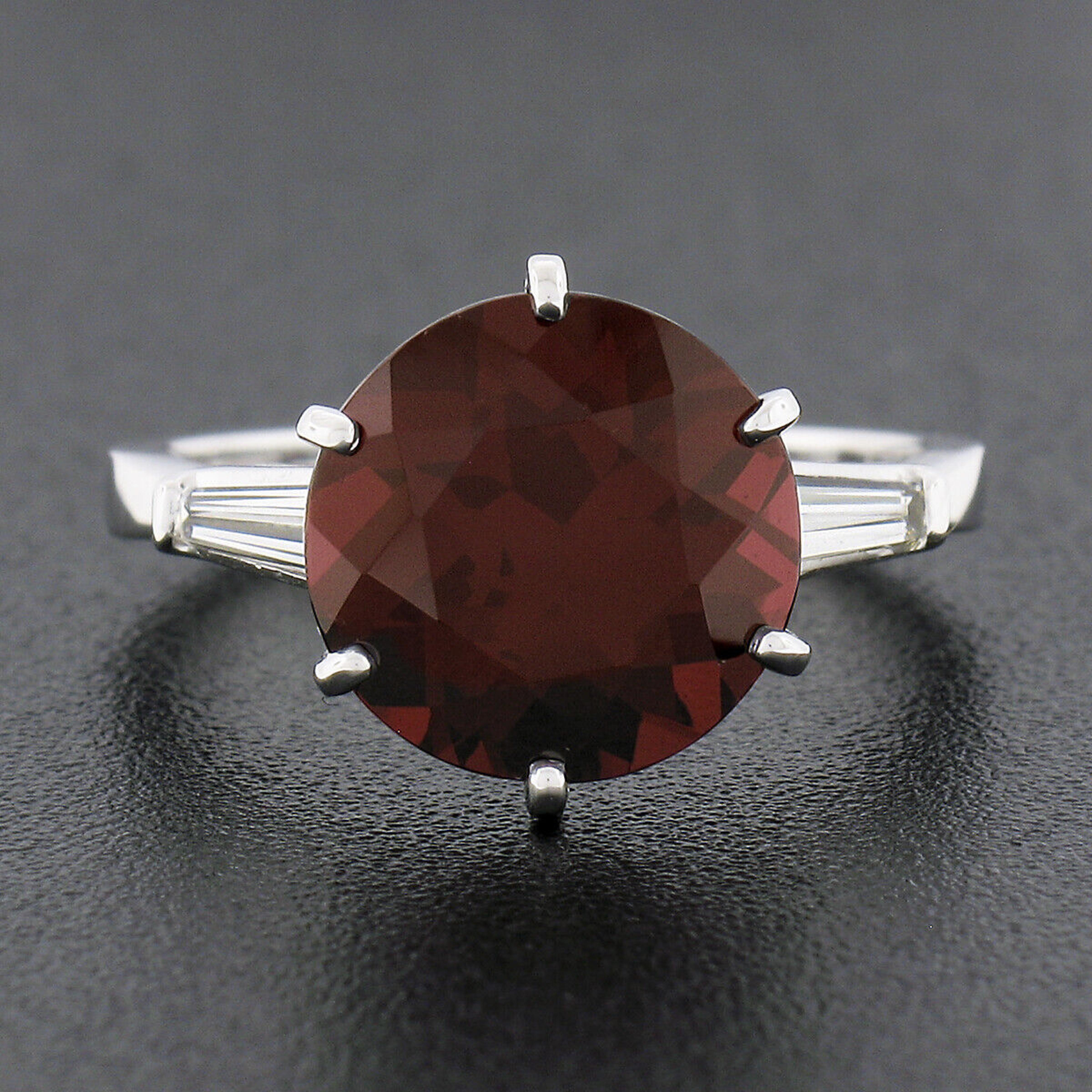 Here we have a truly breathtaking vintage ring crafted from solid platinum featuring an elegant 3-stone style that carries an absolutely incredible round brilliant cut garnet 6-prong set at the center with tapered baguette diamonds neatly channel