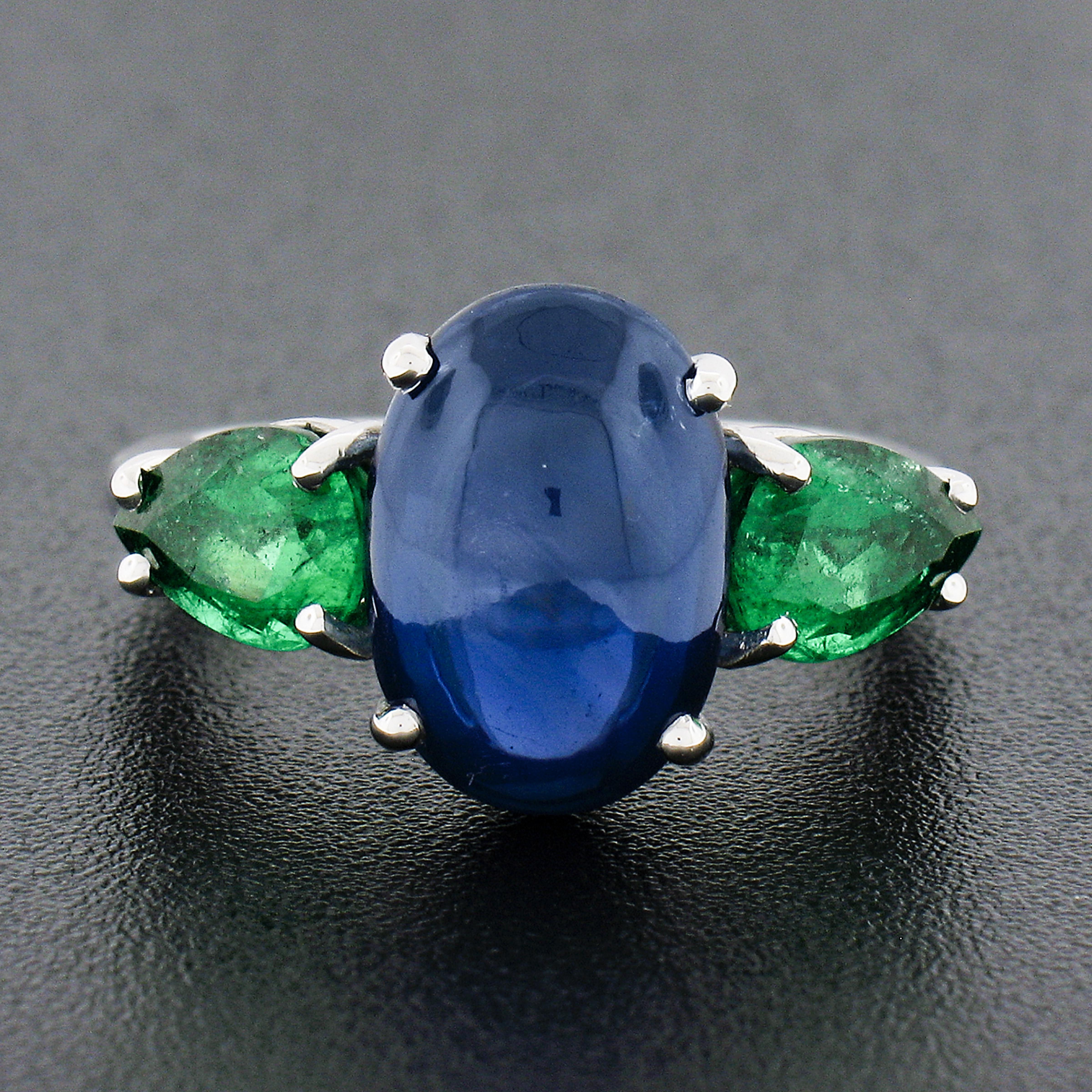 You are looking at an absolutely breathtaking, vintage, sapphire and emerald three stone ring that is very well crafted in solid platinum. The ring features a beautiful, GIA certified, elongated oval cabochon cut sapphire solitaire neatly prong set