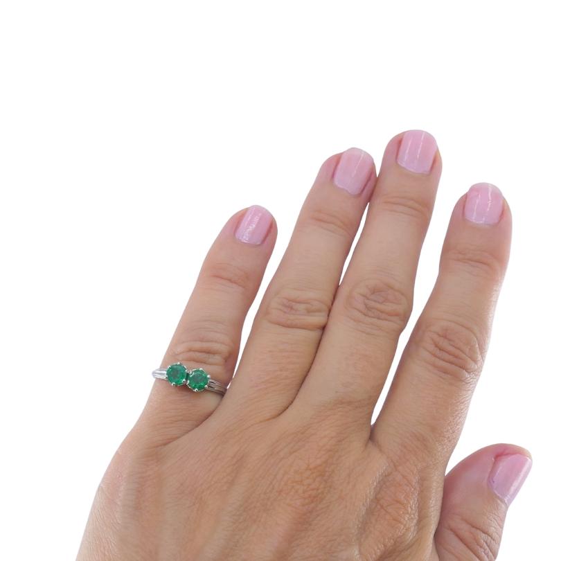 This vintage platinum ring features two round green emeralds set side by side in classic prong baskets, creating a balanced two‑stone design that symbolizes partnership and connection. Each emerald is held in a multi‑prong setting. The shoulders are