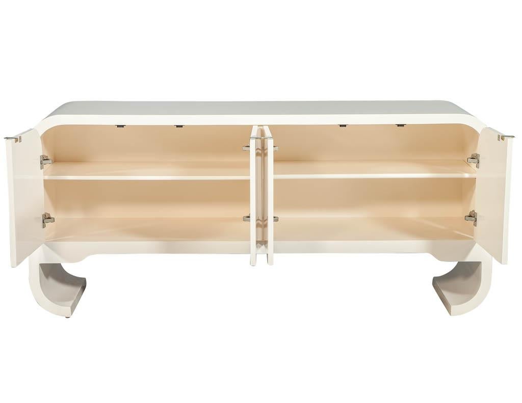 Vintage polished waterfall buffet credenza. Featuring sleek transitional design, curved waterfall ends, and beautiful accented brass hardware. Finished in a off white polished lacquer with a faint blush undertone.