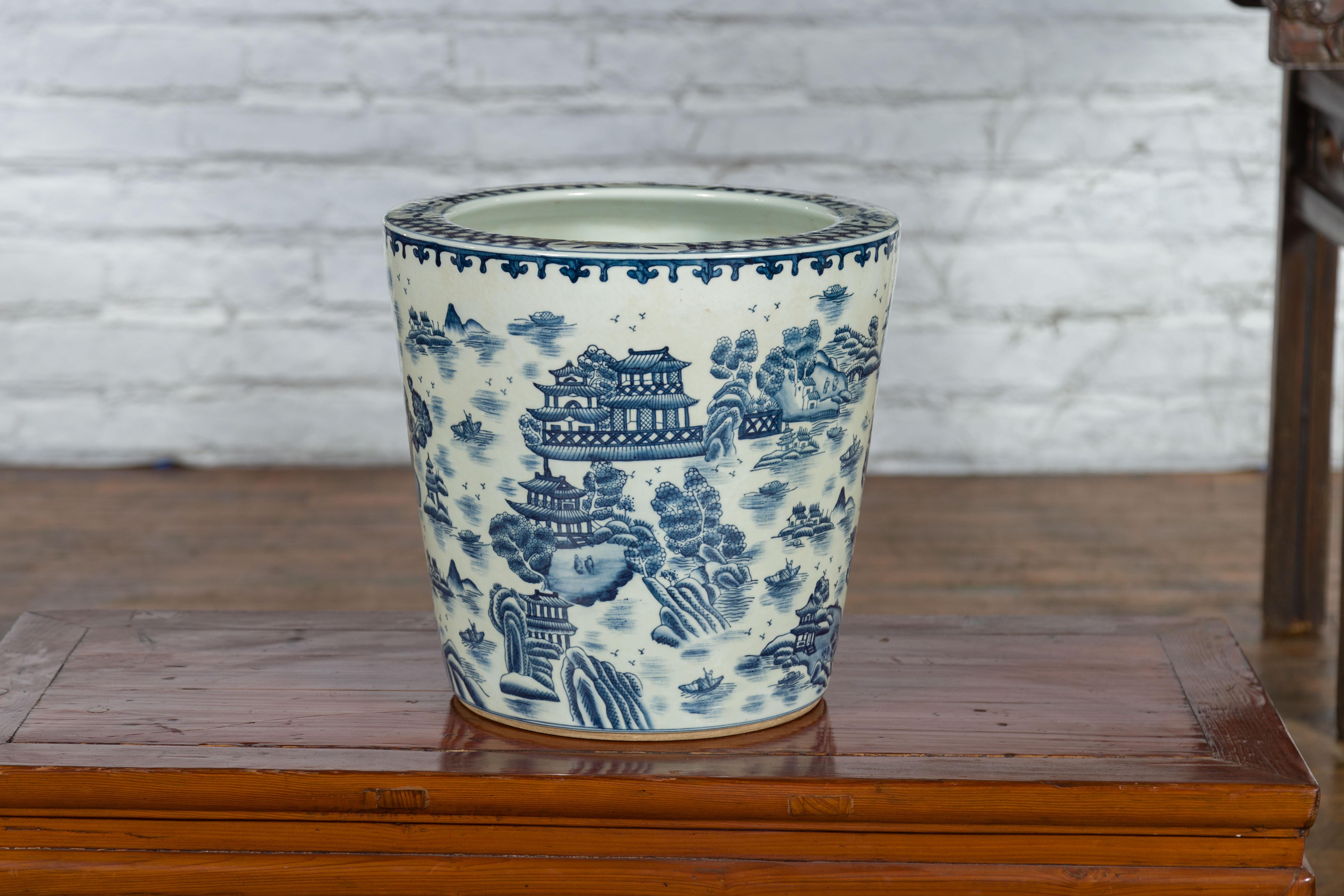 A Chinese vintage porcelain cache-pot planter from the mid 20th century with hand-painted blue and white décor depicting landscapes, architectures and figures in boats patterns. Created in China during the midcentury period, this porcelain cache-pot