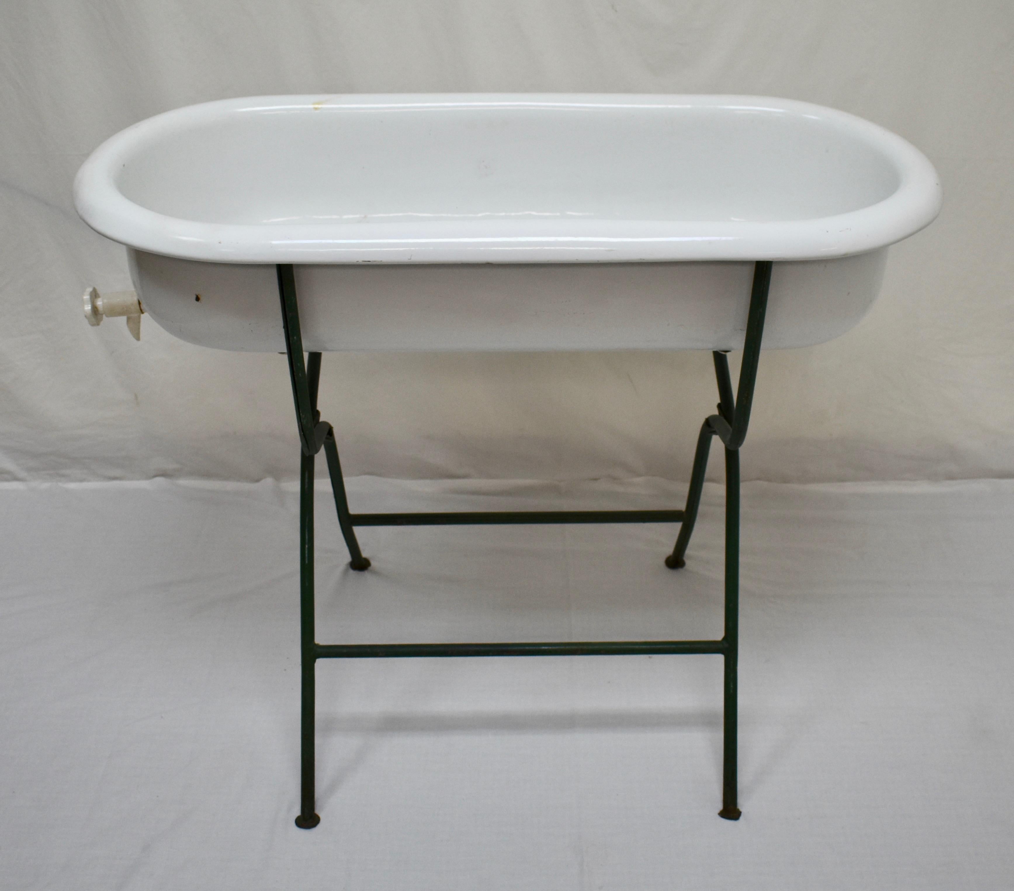 This is a vintage porcelain enamel baby bath on its original wrought iron folding stand. This is a particularly nice example, in that its stand is painted a deep hunter green and the plastic drain tap, an old replacement, is in good working order.