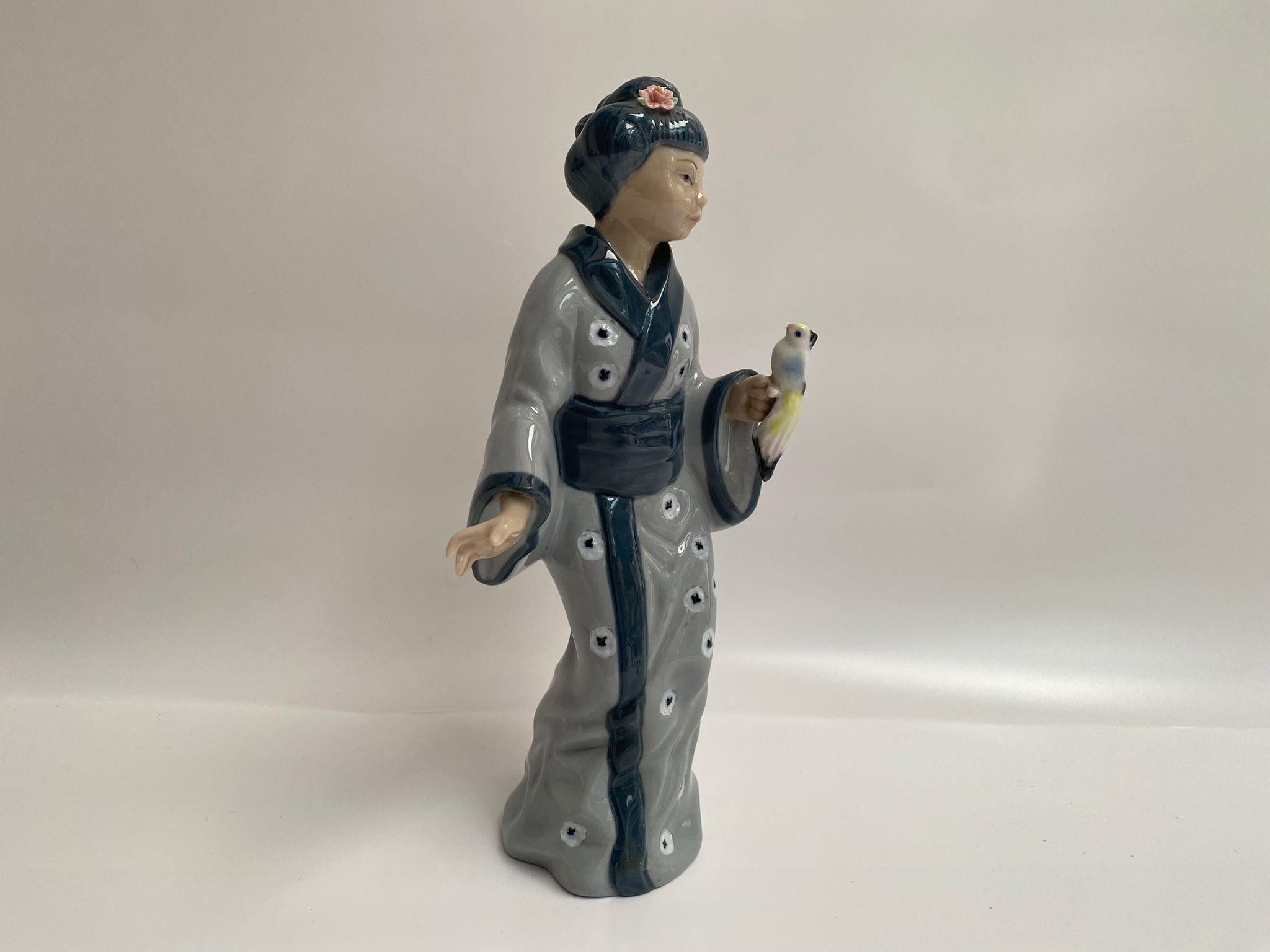 La figurita Geisha de Casades es una exquisita pieza de porcelana de la década de 1980, elaborada en elegantes tonos grises y azules. La geisha va vestida con un kimono tradicional adornado con motivos florales, y un delicado pájaro posado en su