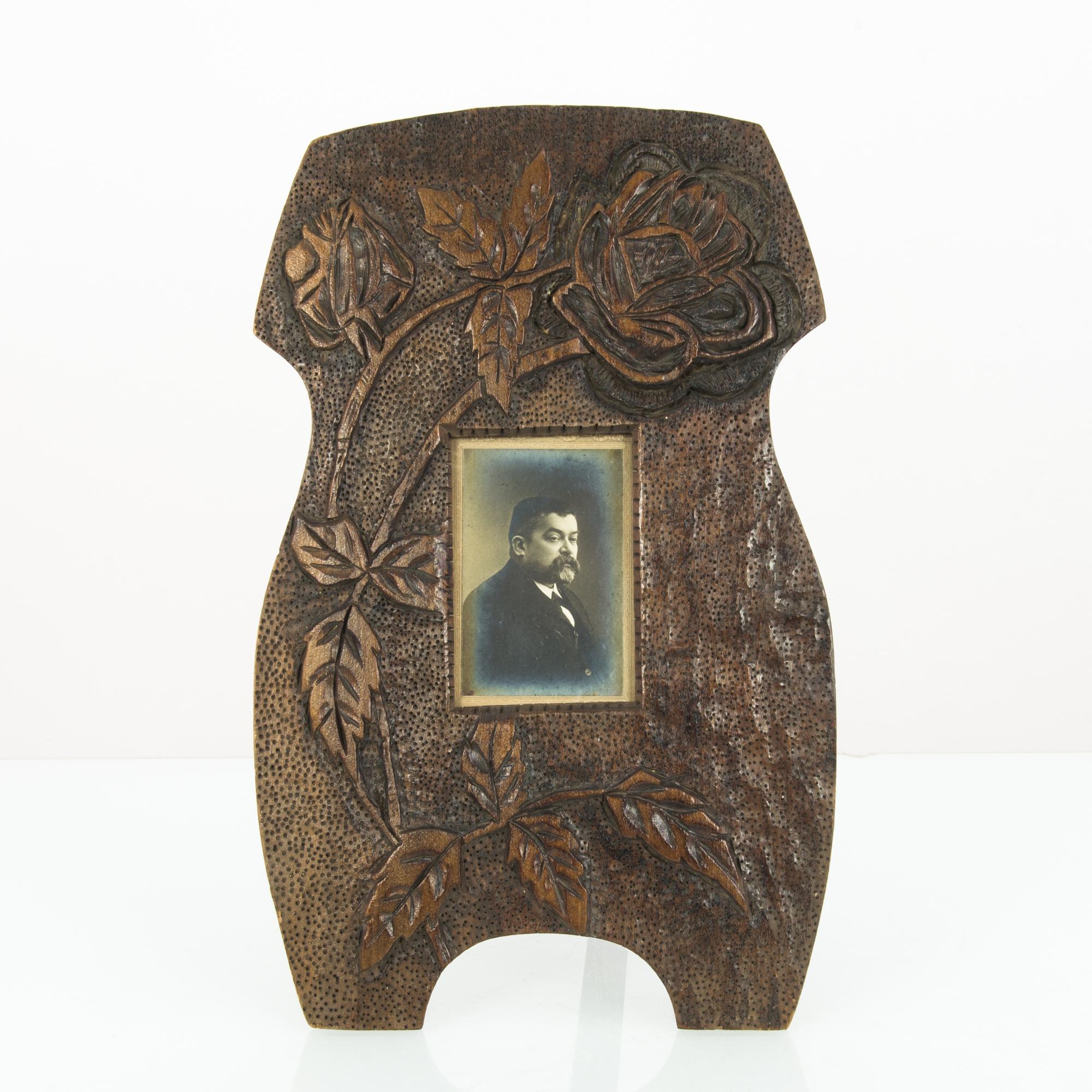 A photographic portrait in a carved wooden frame from Belgium, circa 1920. The frame is ample, while the print is small: the photograph is positioned under the sweep of a carved rose in full bloom. The edges of the frame curve in a sinuous,