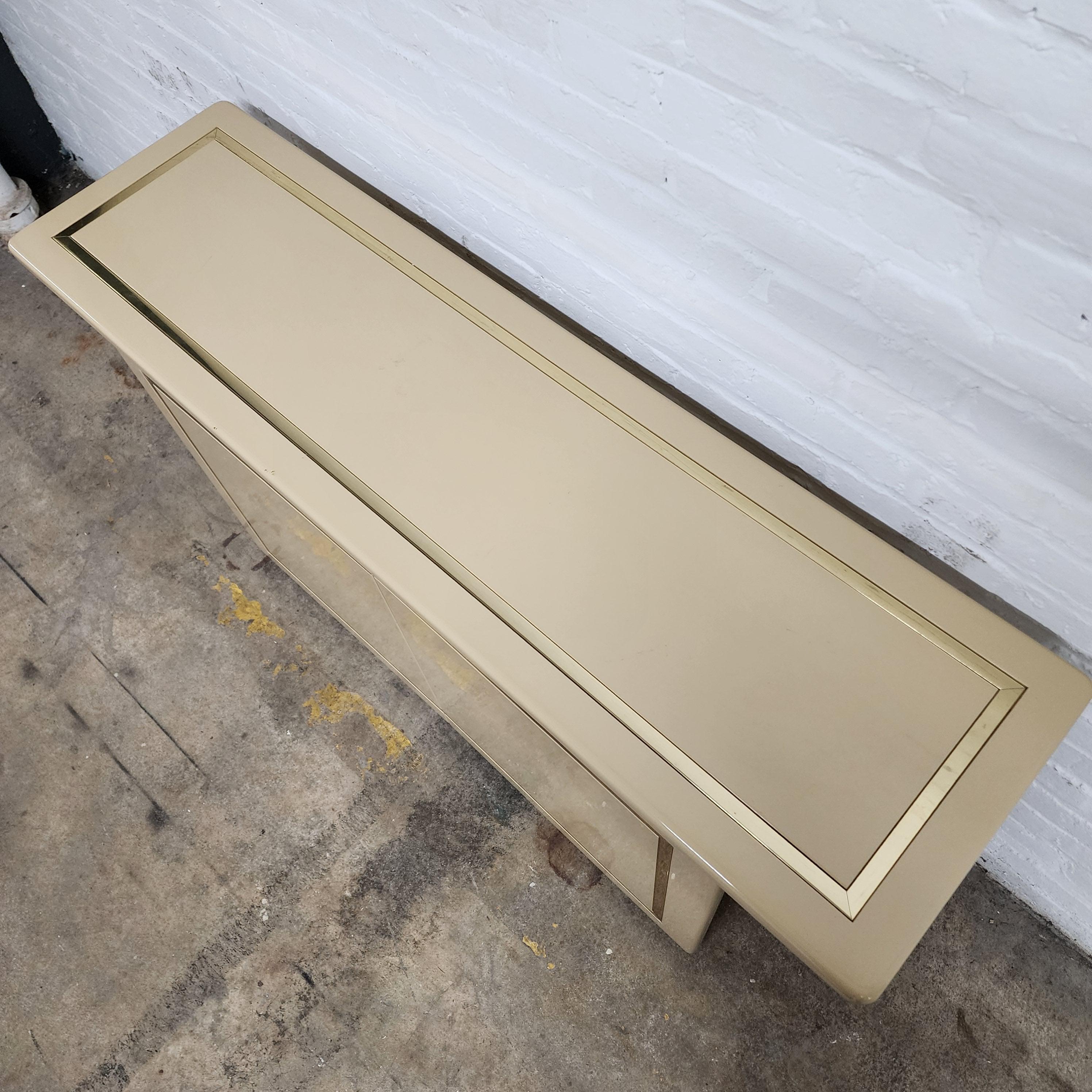 This post-modern cream lacuqer and brass credenza by Rougier is a sleek and stylish piece of furniture that embodies the eclectic, forward-thinking design sensibilities of the post-modern era. The credenza features a cream-colored body that is both