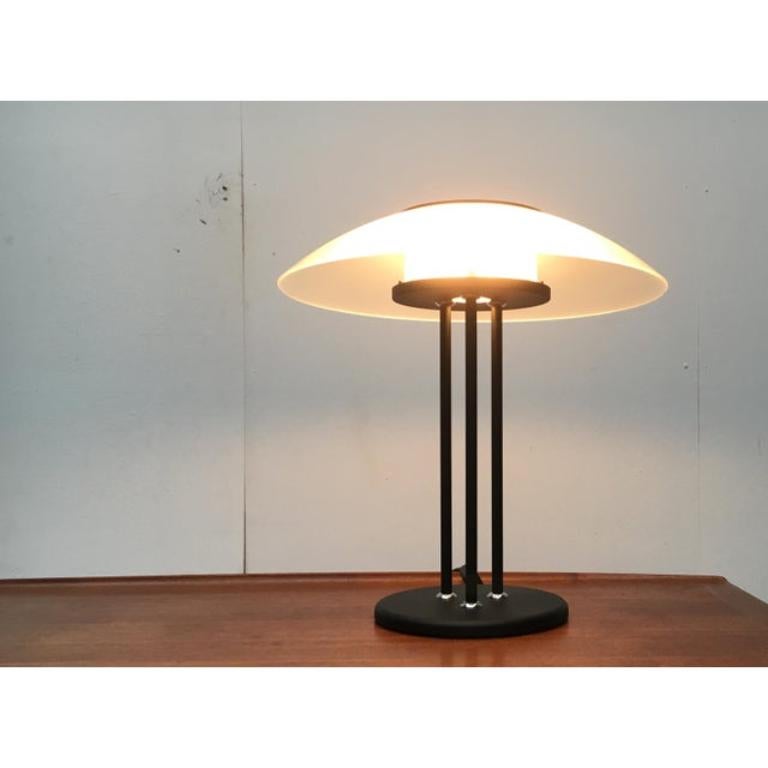 Lámpara de mesa grande en lenguaje de diseño postmoderno.

Imponente lámpara de sobremesa con gran difusor de cristal. La base de la lámpara se resuelve en tres soportes que se asientan sobre una sencilla base de chapa. La transición se establece en