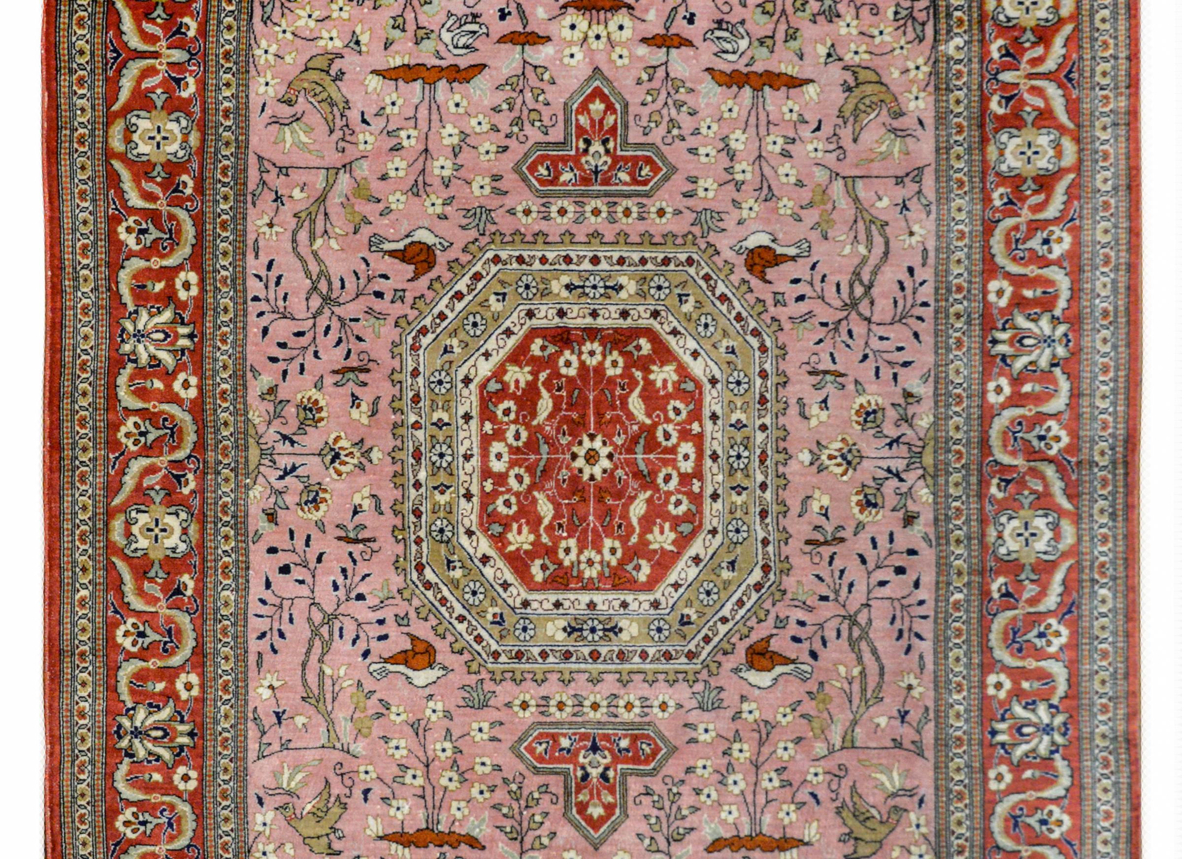A beautiful vintage Persian Qum silk rug with an eight sided medallion with a floral pattern set amidst a field of myriad flowers, birds, and deer, all woven in brown, white, and crimson, set against a pink background, and surrounded by a complex