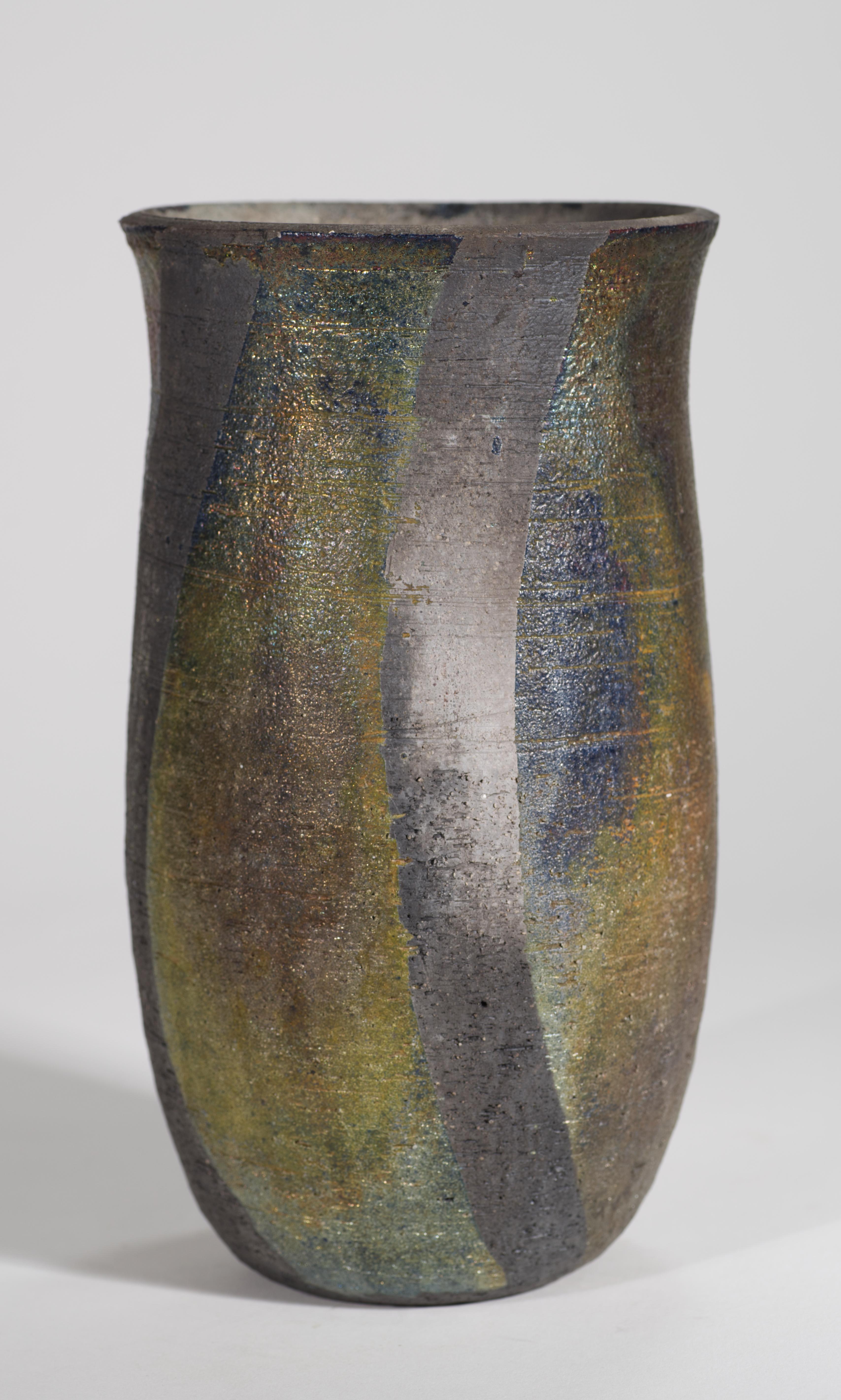 Il vaso scultoreo in ceramica presenta una finitura Raku metallizzata multicolore con toni predominanti di grigio, marrone e verde. Lo smalto complesso e grafico completa la forma minimale del vaso, guidando l'occhio lungo le linee del pezzo.

Il