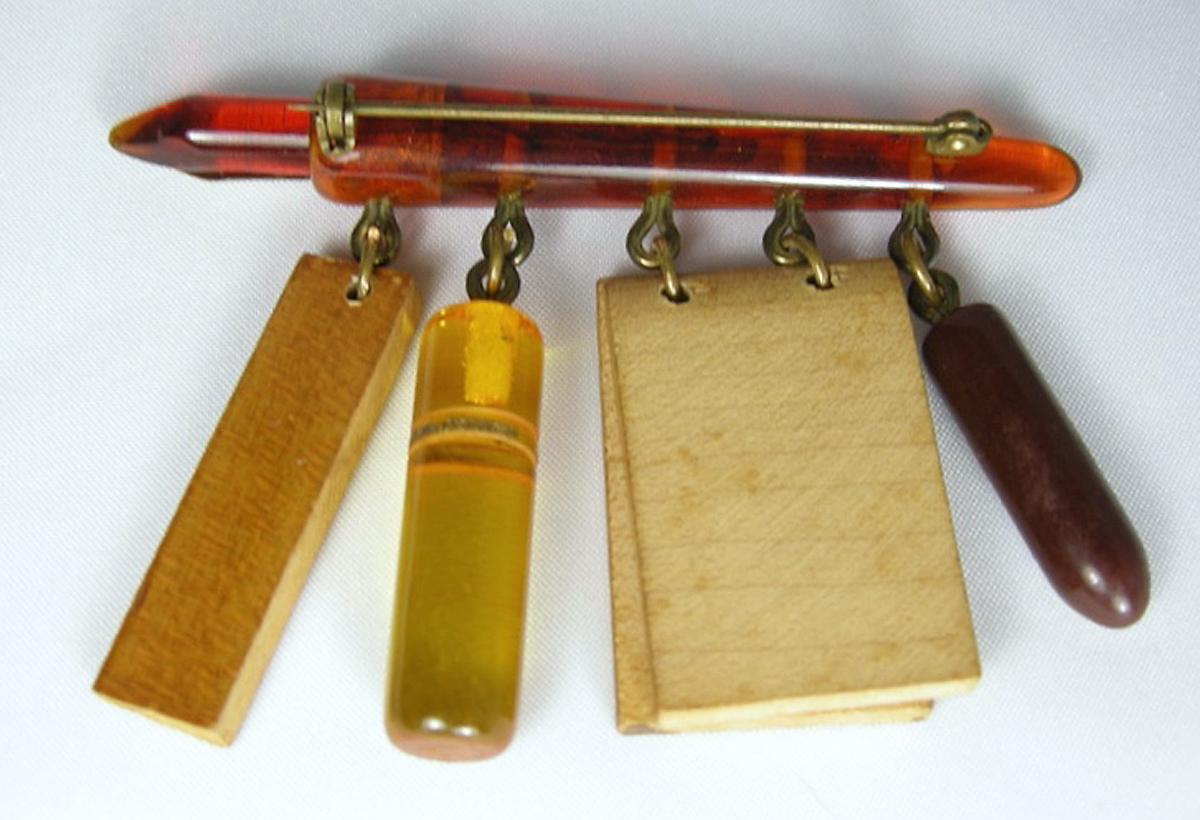 This rare 1930s brooch comes from the school days Bakelite collection. It has an old-fashioned fountain pen made of apple juice Bakelite with a gold tone nib.  There are four dangling items representing school days made of wood and Bakelite.  It