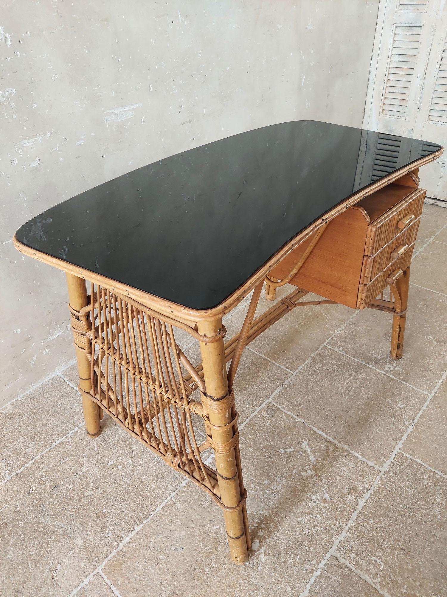 Bureau vintage en rotin et bambou. Une jolie table d'écriture en rotin avec trois tiroirs et un plateau en verre noir légèrement arrondi.

h 76 x l 110 x 60 cm