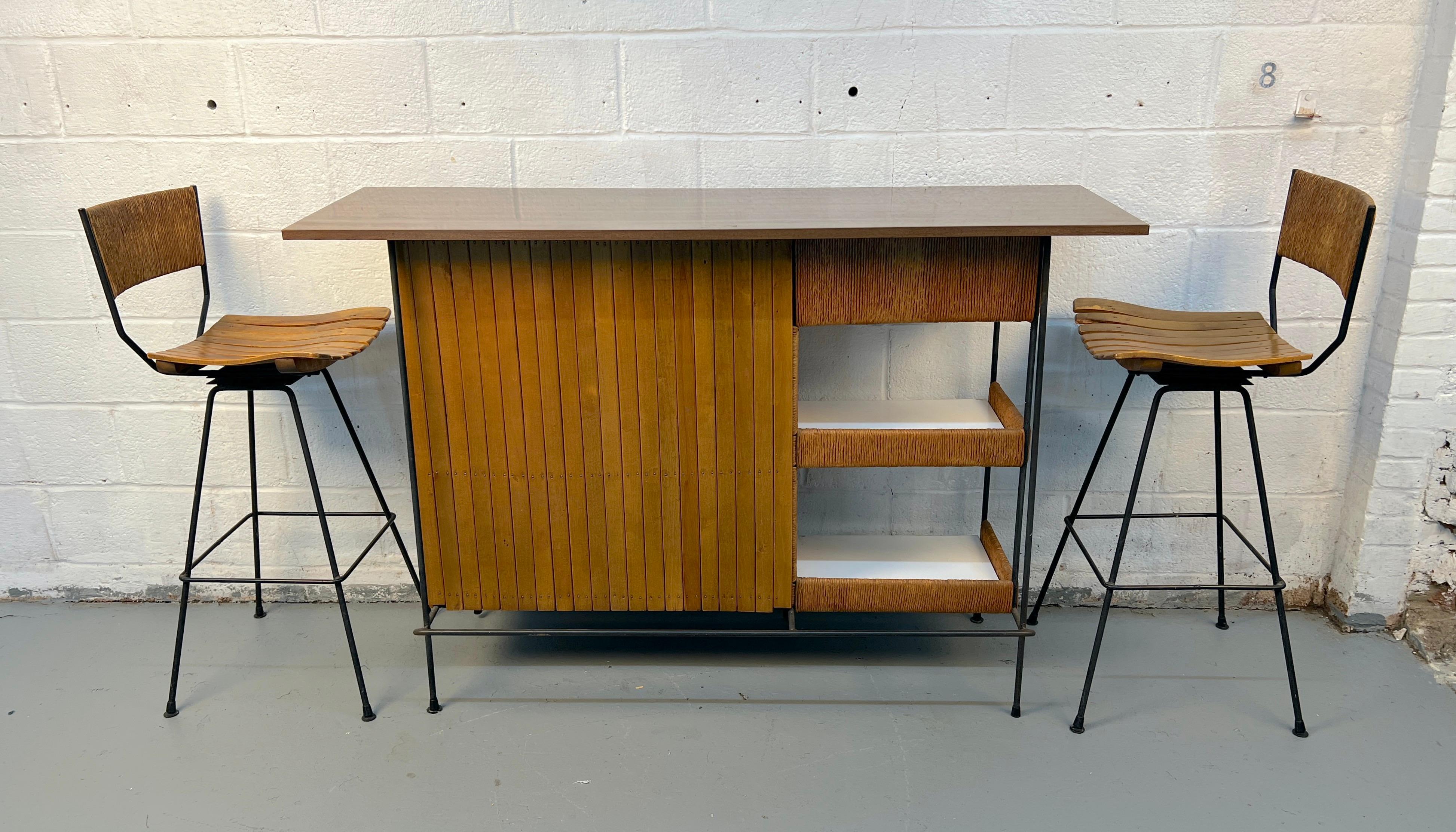 Elegant ensemble de bar du milieu du siècle, avec une façade en lattes de bois, un plateau en stratifié élégant et (2) tabourets en rotin. Les étagères ouvertes offrent un rangement pratique tout en conservant un profil léger et moderne. Deux
