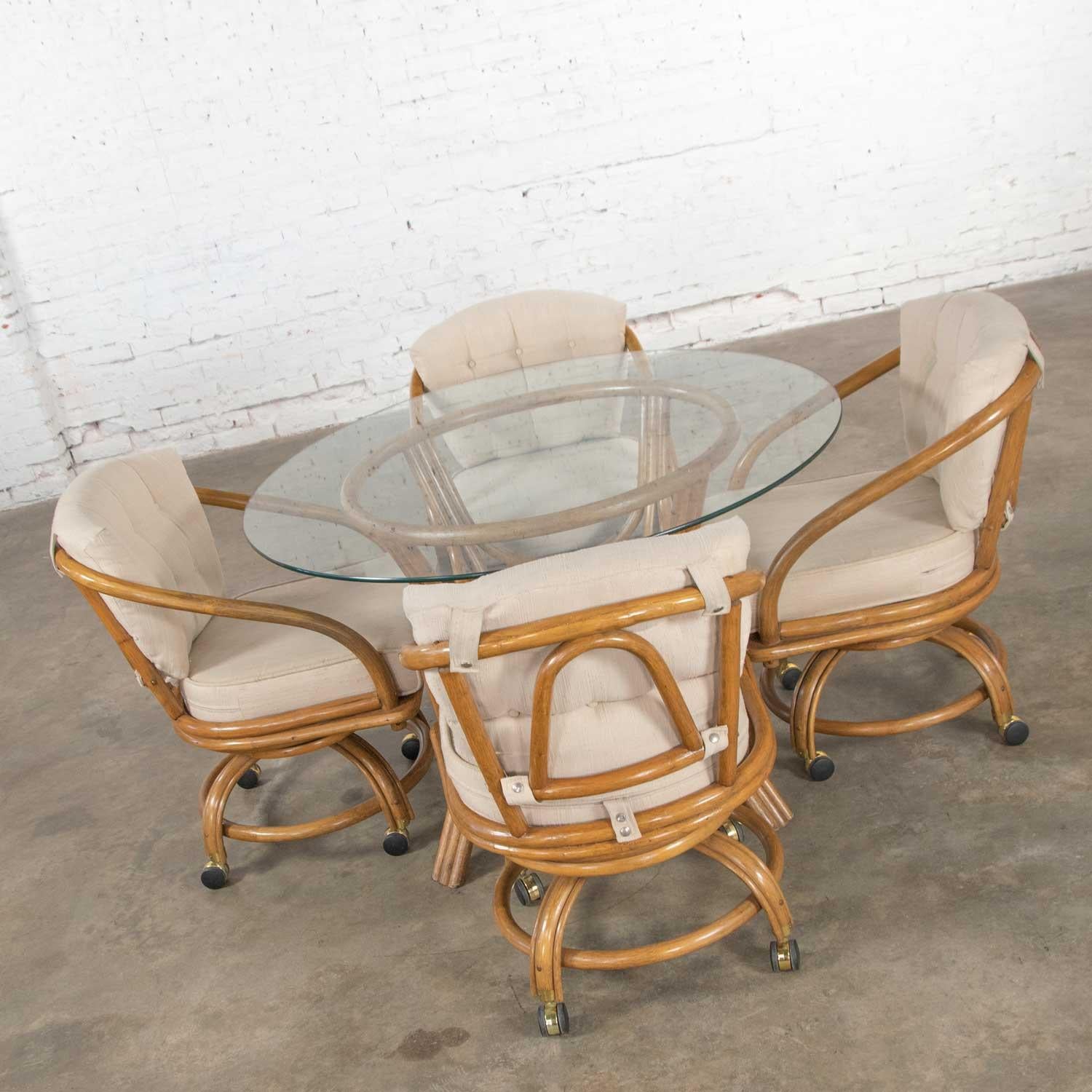 Handsome game table or dining table set including a round glass top rattan base table and four (4) swivel rolling rattan chairs with oatmeal colored upholstered removable seat and back cushions. They are in wonderful vintage condition. The cushions