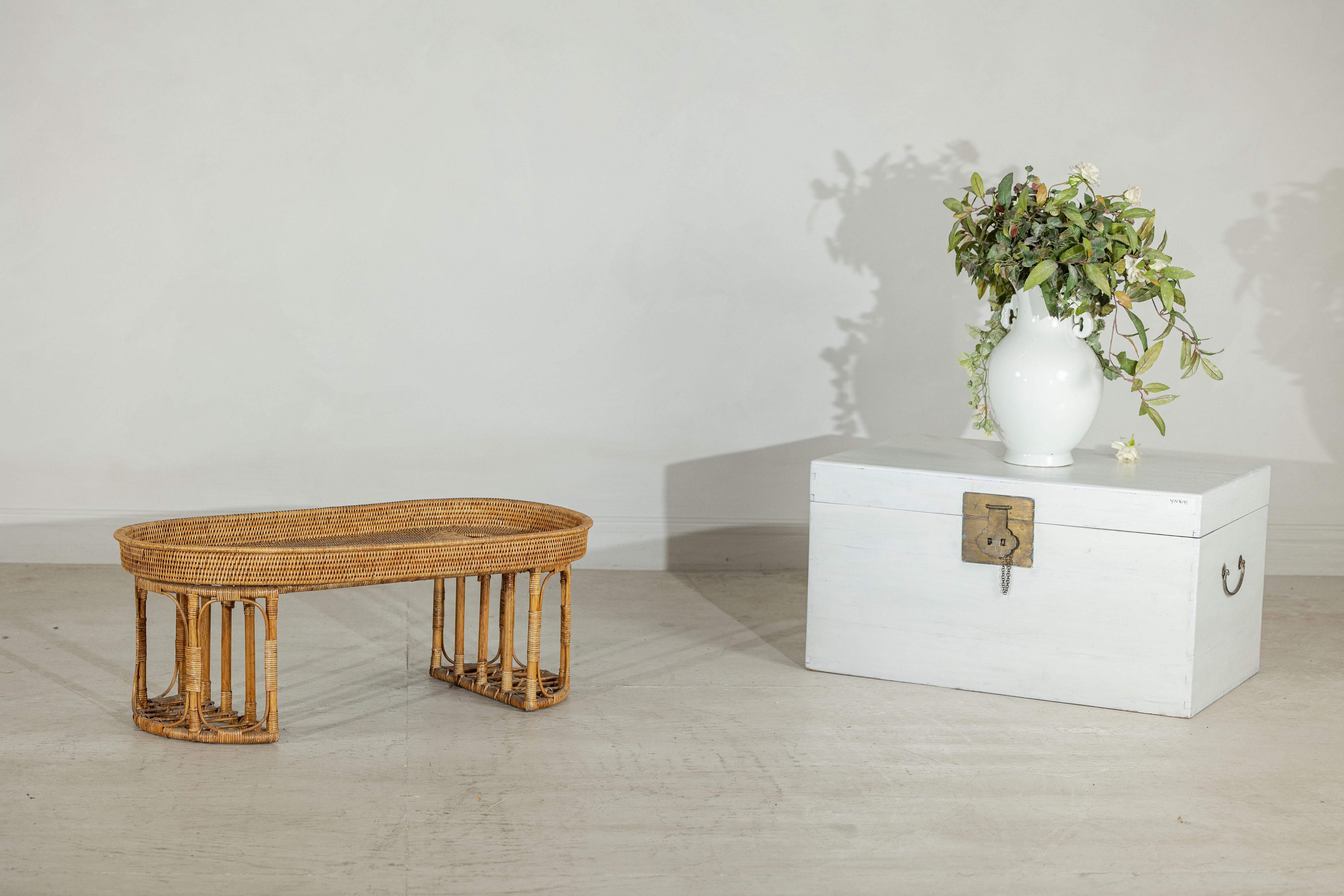 Questo tavolo in rattan vintage offre una presenza versatile e affascinante, adatta sia come tavolino basso che come grande vassoio da portata. Il bordo ovale rialzato è finemente intrecciato e si trova sopra un'ariosa base traforata formata da pali
