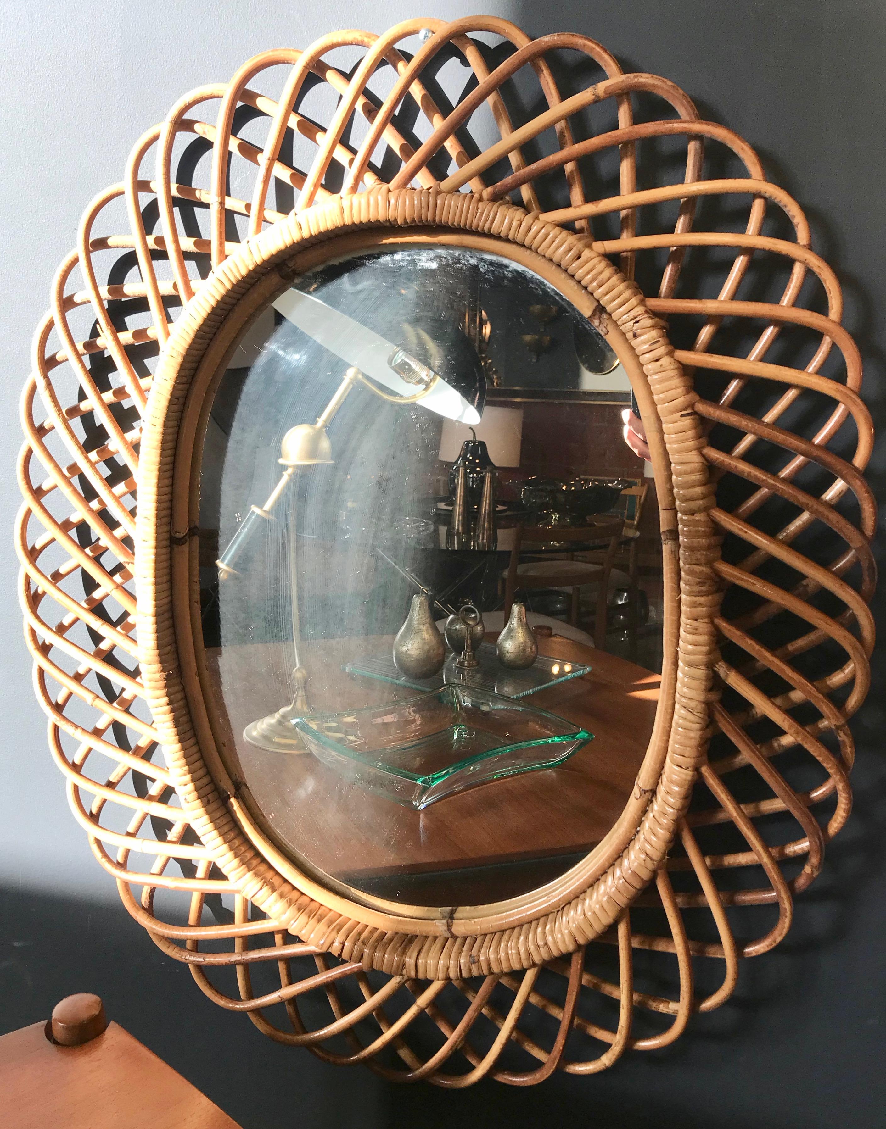 Italian rattan oval wall mirror (circa 1960s) in the style of Franco Albini. This mirror has a complex weave of rattan in a series of horseshoe-shaped projections on the frame edge. There is a graceful, aged patina on the mirror frame and the glass