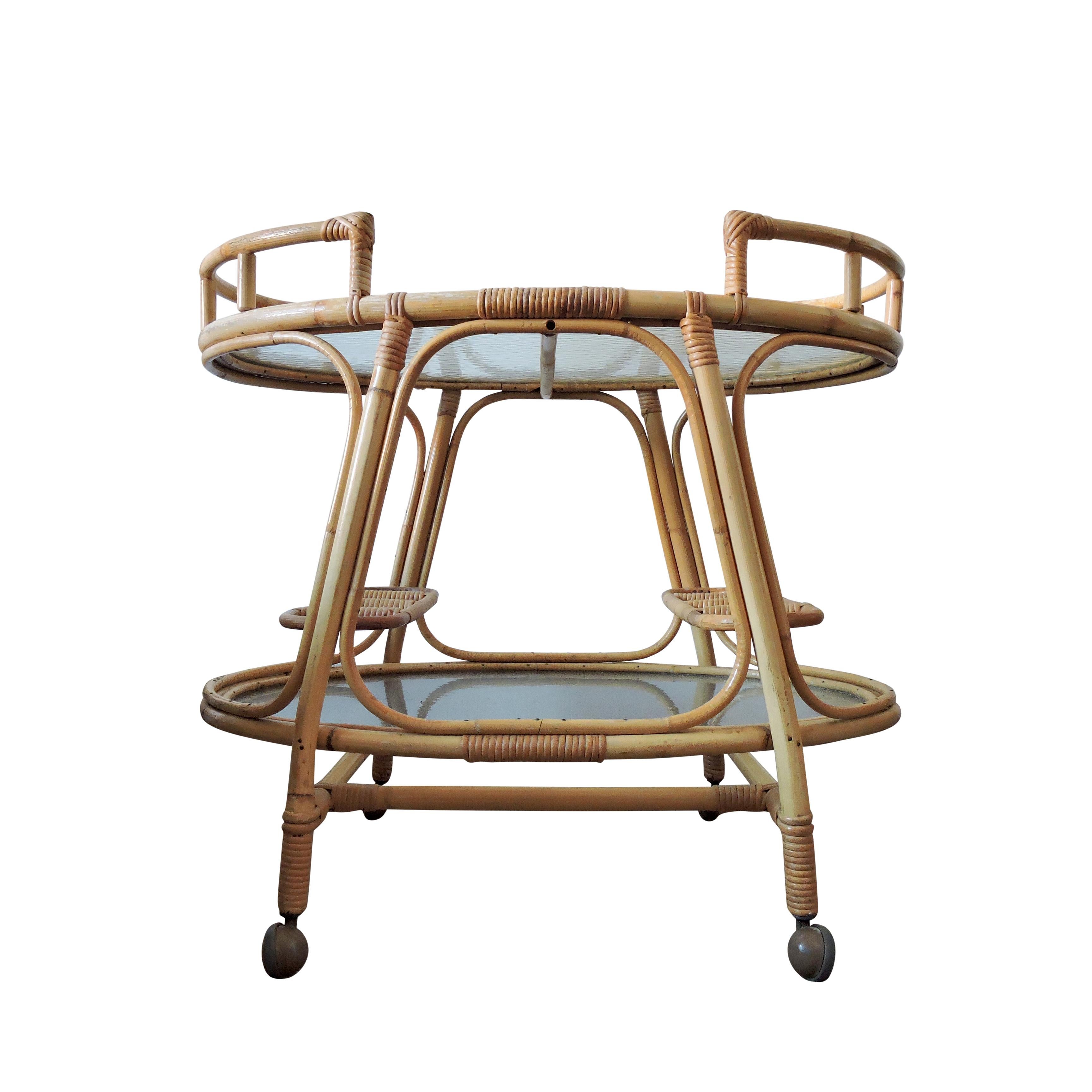 A 1960s oval rattan bamboo drinks trolley with industrial glass shelves and two integrated bottle holders on lower shelf.
        