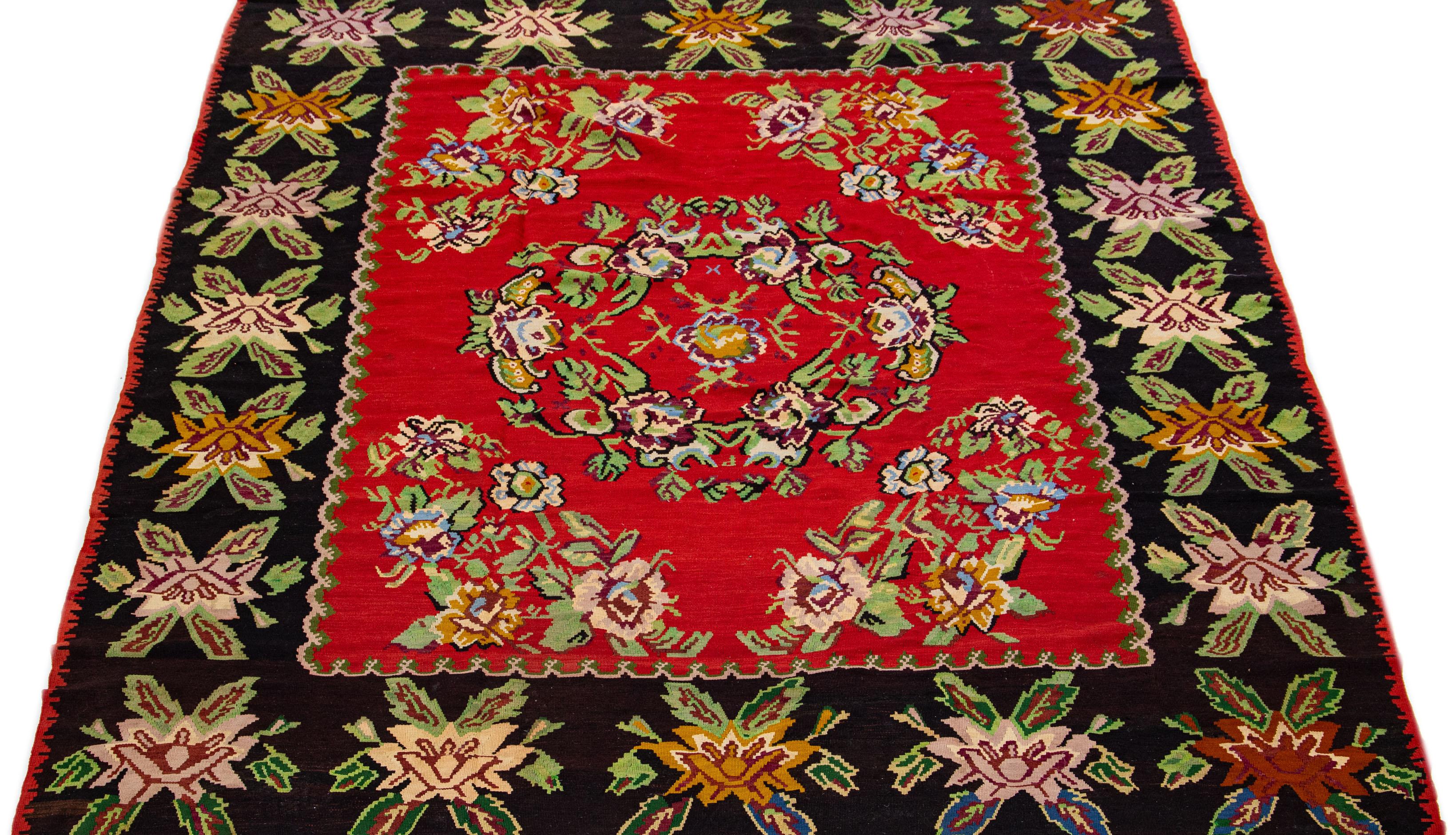 This exquisite Bessarabian Style Kilim rug features a striking floral pattern showcased against a rich red backdrop. The charming accents of green, blue, pink, and black add a delightful touch of color to this meticulously hand-knotted
