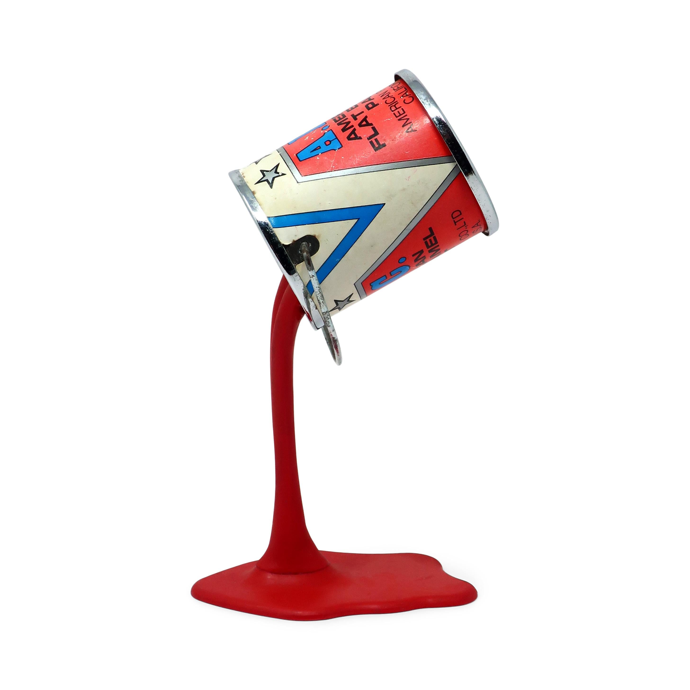 Una hucha/escultura en forma de lata de pintura de los años 80 perfectamente Pop Art.  Una lata metálica de pintura con una ranura para monedas del tamaño de un cuarto en su parte inferior parece desafiar a la gravedad al descansar sobre un chorro