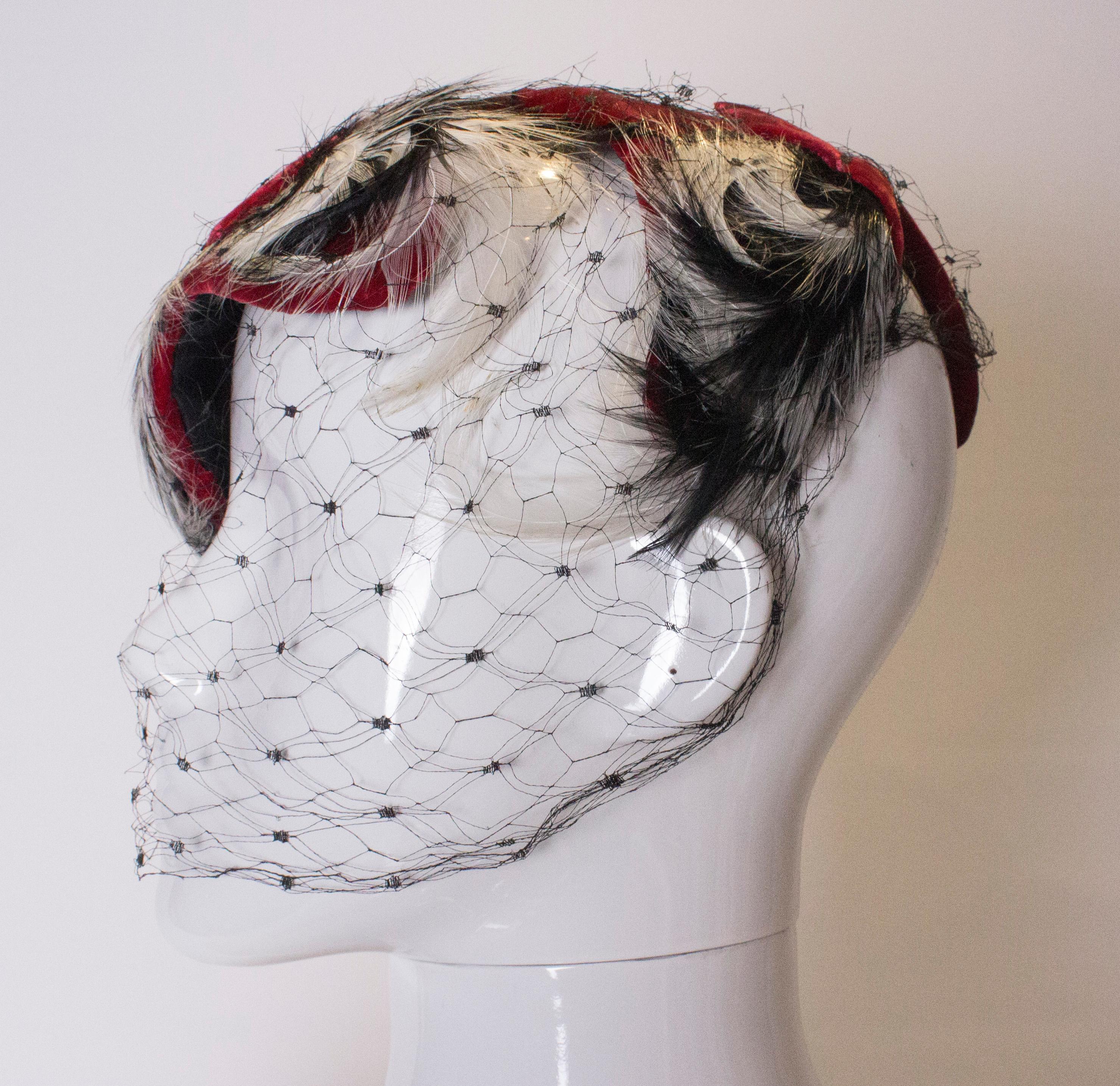 A head turning hat from the 1950s. The crown of the hat is a pretty red velvet with two decorative bows.The pretty feathers add a colour contrast and the net covers the face.