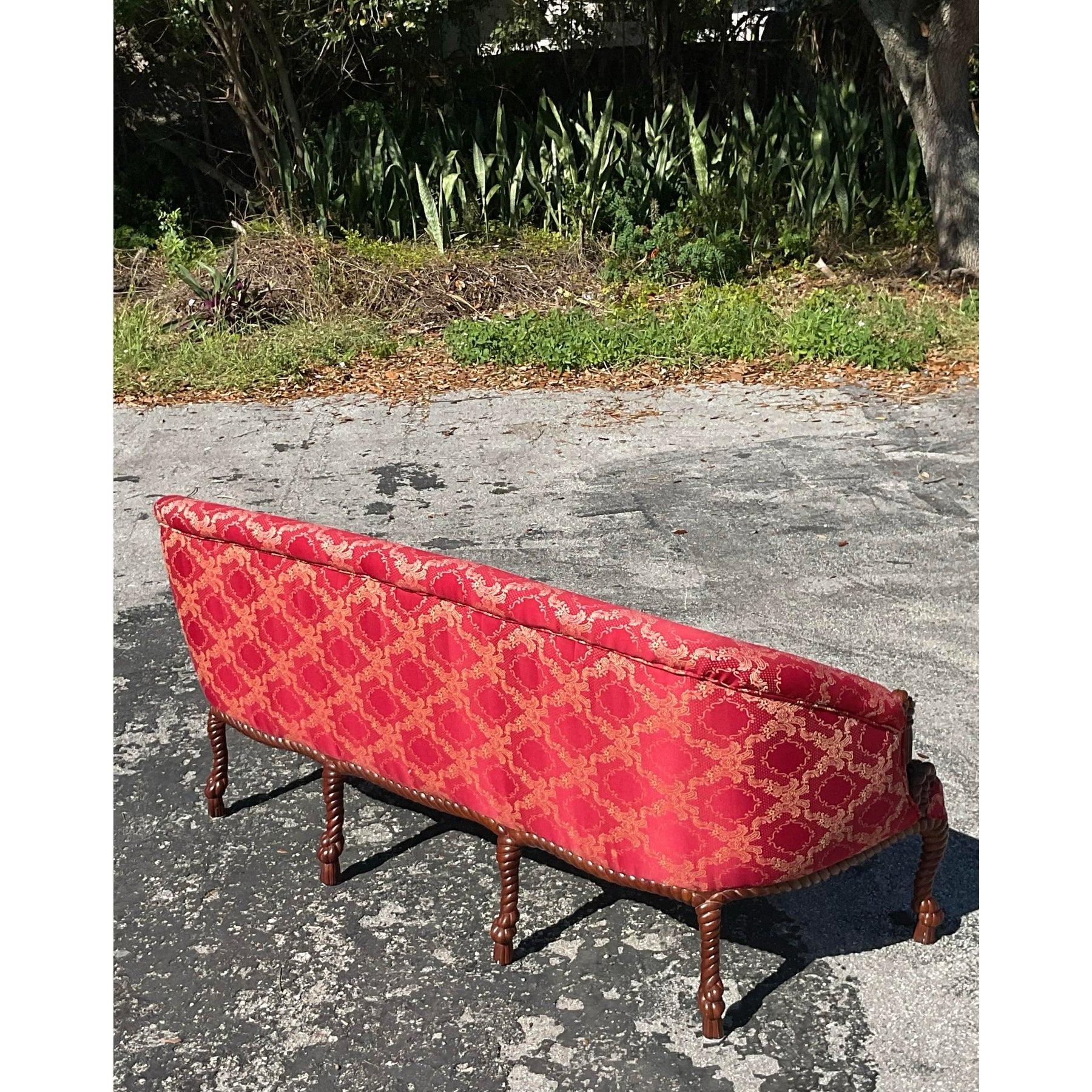 Este impresionante sofá vintage de estilo Regencia destila sofisticación atemporal con su marco de madera de nudos y cuerdas bellamente tallado. Tapizada en un llamativo tejido adamascado rojo y dorado, esta elegante pieza exhibe una artesanía