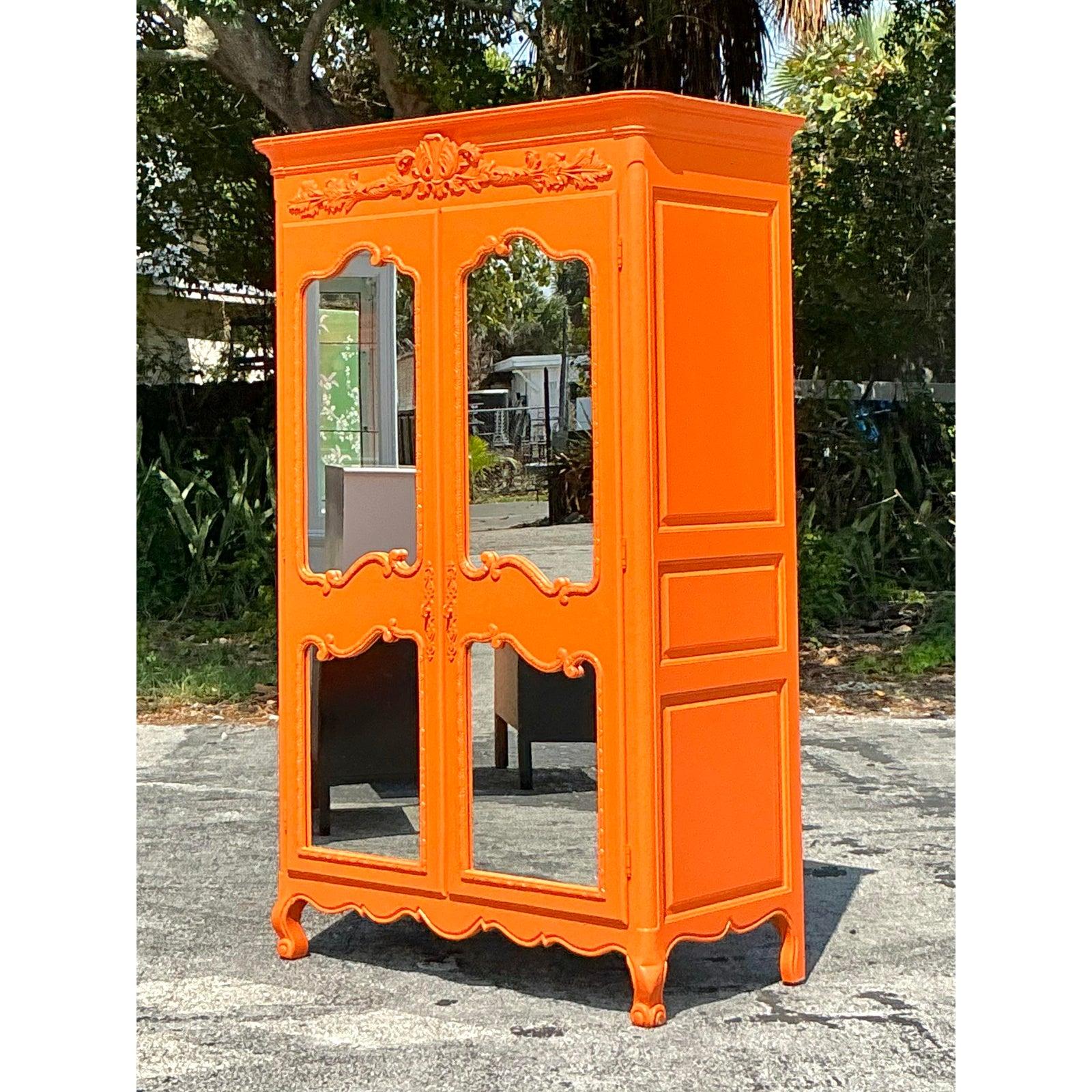 Setzen Sie ein gewagtes Design-Statement mit diesem auffälligen, lackierten Vintage-Regency-Schrank. Das in leuchtendem Orange lackierte Möbelstück besticht durch florale Schnitzereien, geschwungene Kanten und verspiegelte Türen im französischen