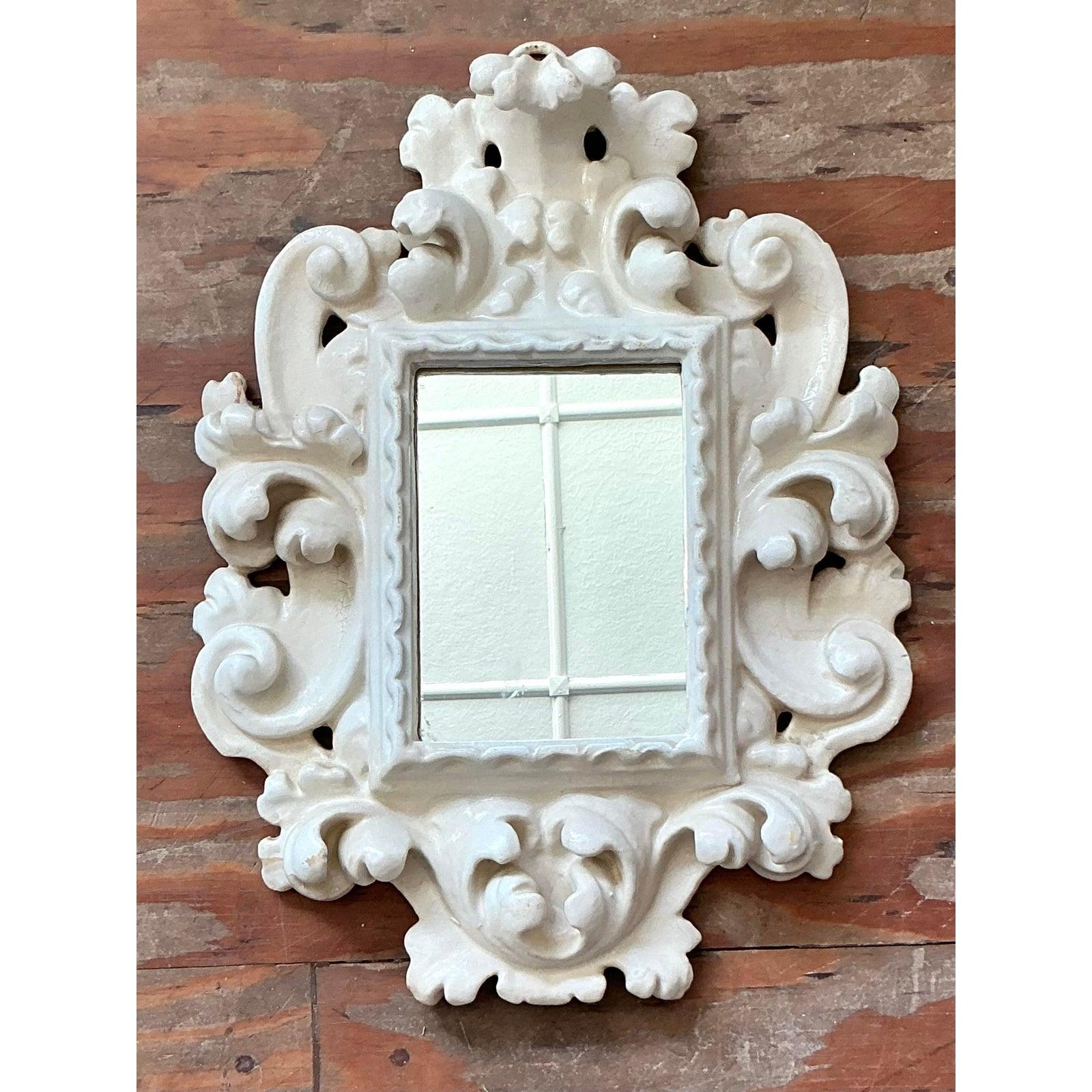 Ce charmant petit miroir mural capture la courbure extravagante et la richesse décorative du style Rococo, réinterprété pour une esthétique vintage Hollywood Regency ou Grandmillennial. Le cadre est moulé de façon complexe, probablement en résine ou