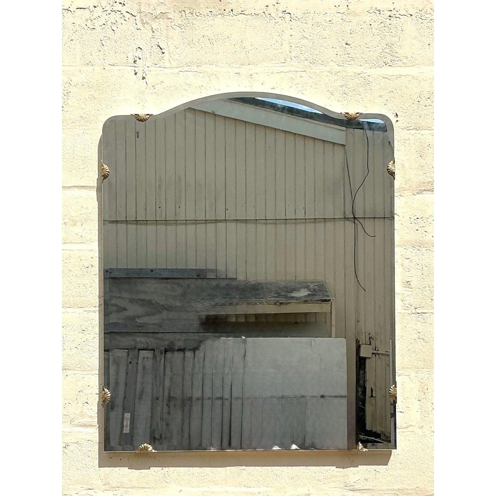Este exquisito espejo vintage de la Regencia presenta un elegante diseño en forma de escudo con la parte superior graciosamente curvada, adornada con decorativos adornos dorados en las esquinas. El cristal biselado realza su refinada estética,