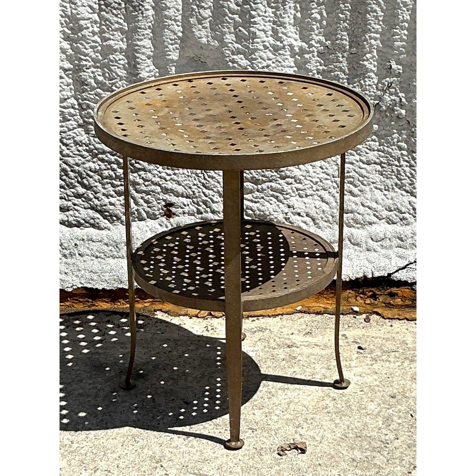 Cette table d'appoint vintage de style Regency est un mélange charmant de fonctionnalité et d'artisanat artistique. Dotée d'un plateau rond en métal perforé et d'une étagère inférieure secondaire, cette table offre à la fois style et rangement dans