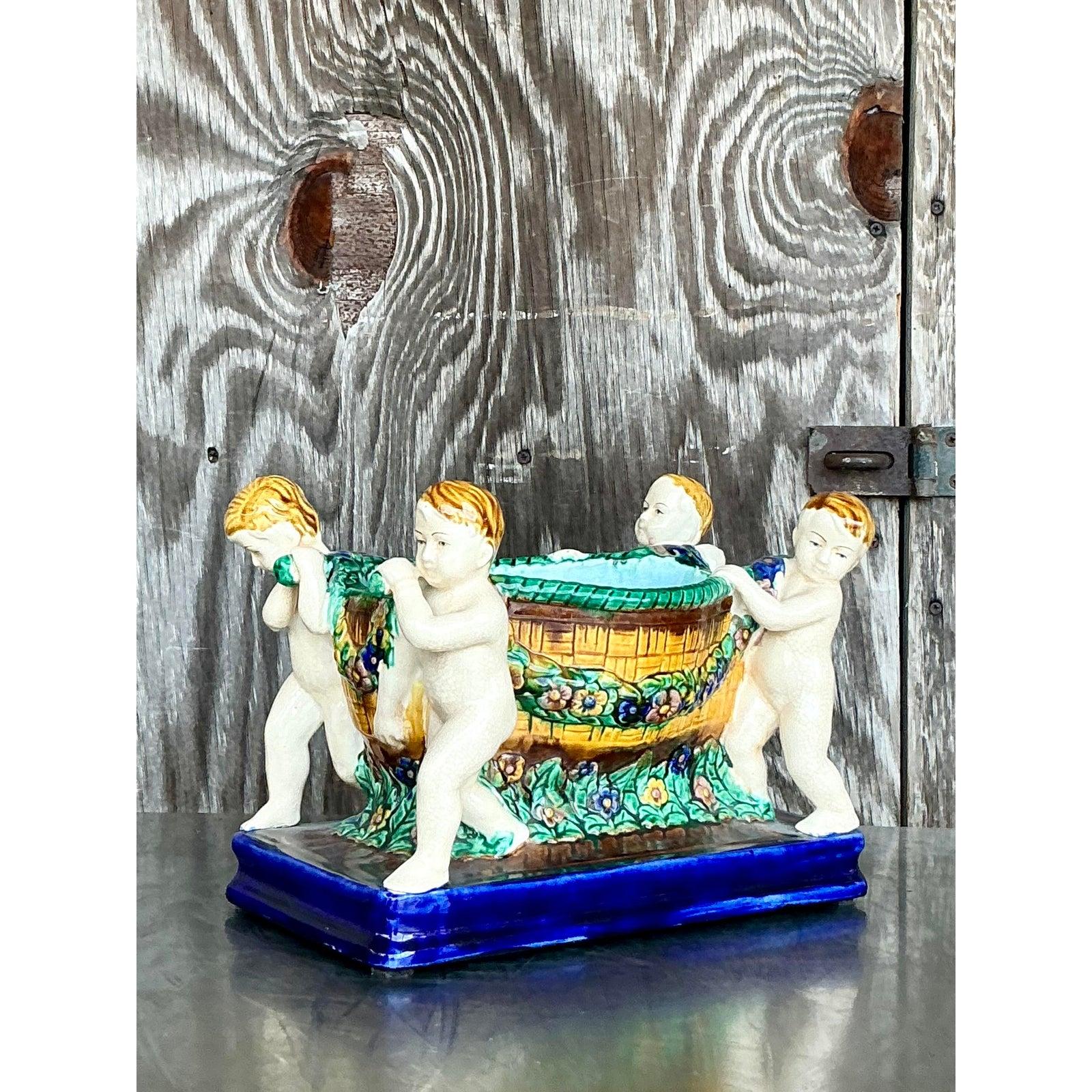 Questo centrotavola in maiolica d'epoca, dal fascino romantico e dalla vivacità artigianale, presenta quattro putti cherubini che sostengono una vasca floreale ricca di dettagli. La scena evoca influenze classiche del Rinascimento con un tocco di