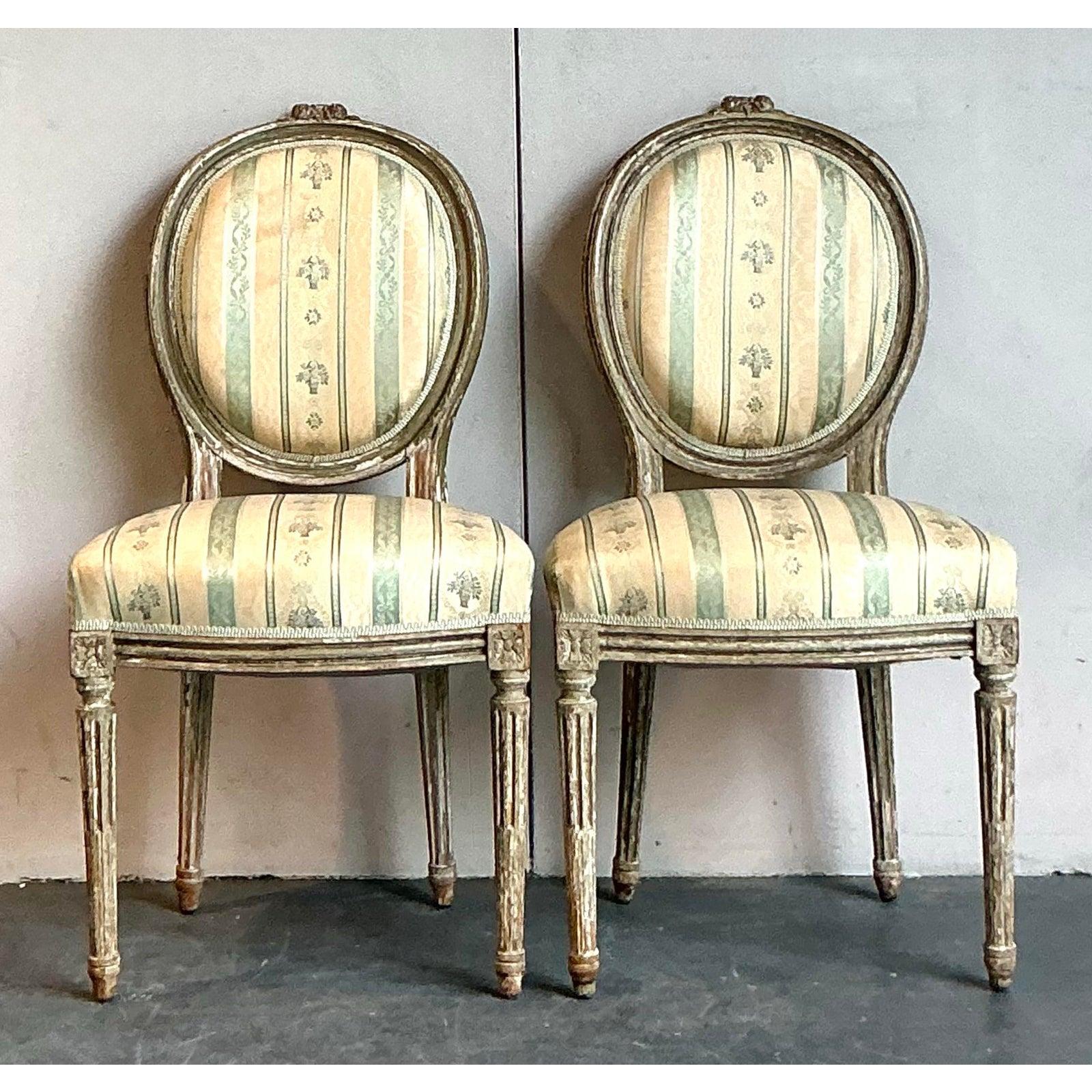 This elegant pair of vintage chairs embodies the sophisticated charm of classic French design with a hint of Regency flair. The beautifully carved wooden frames, featuring a distressed, painted finish, gracefully support oval 