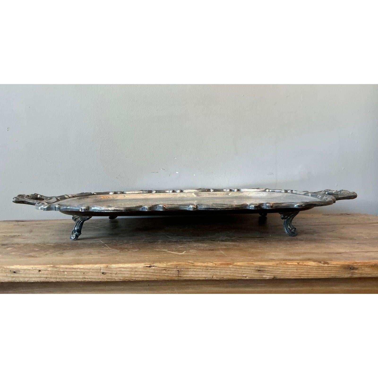 This magnificent vintage serving tray is a true testament to the timeless elegance of Regency-era design. Crafted from polished silverplate, the tray features a stunning oval form with an ornate, raised border and gracefully scrolled handles that