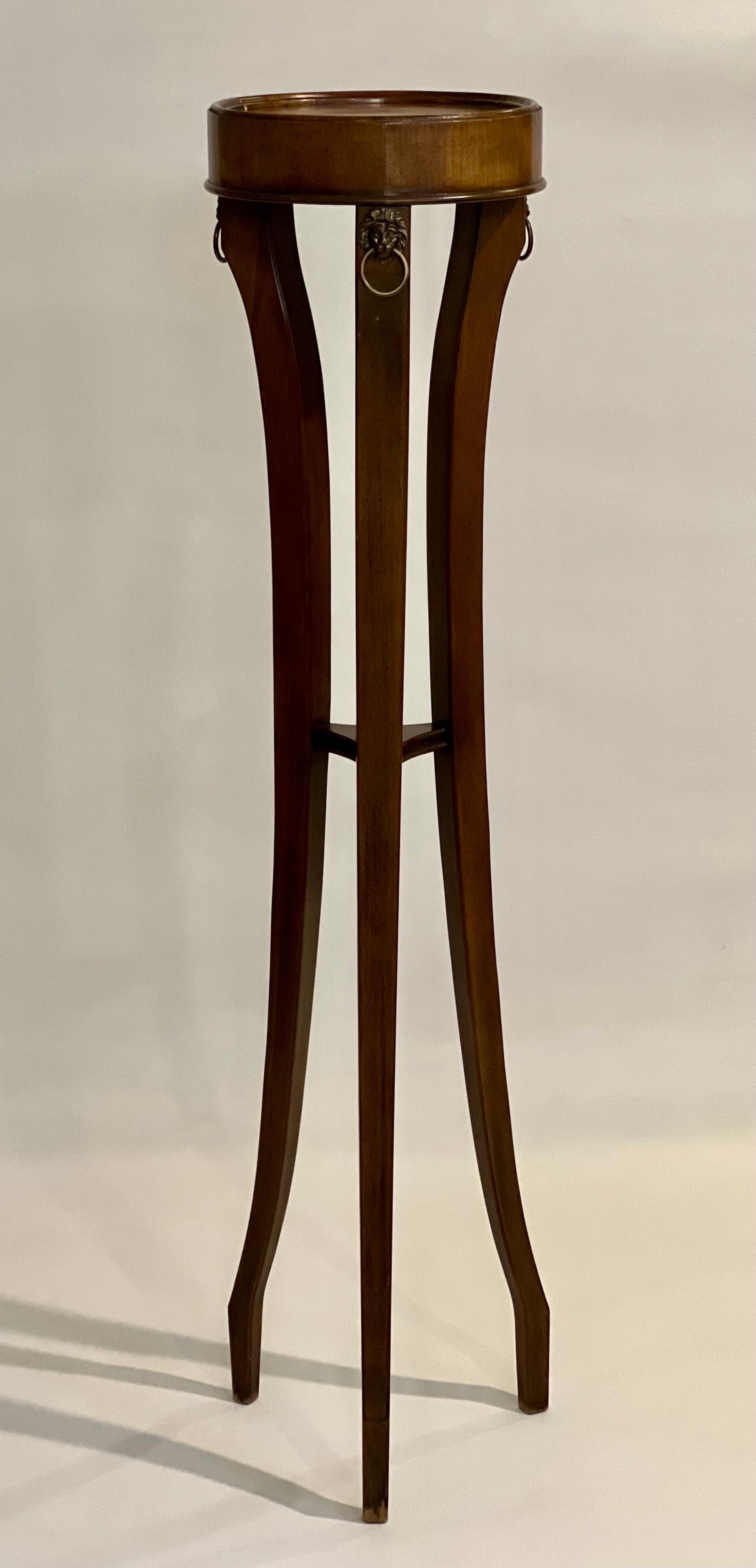 Tall and elegant Regency style walnut sculpture or plant stand, c. 1980.

Stand has a small in-curving lower shelf joined by three shaped legs and brass rings with lion motif at the top. The circular top has a nicely finished, raised rounded edge