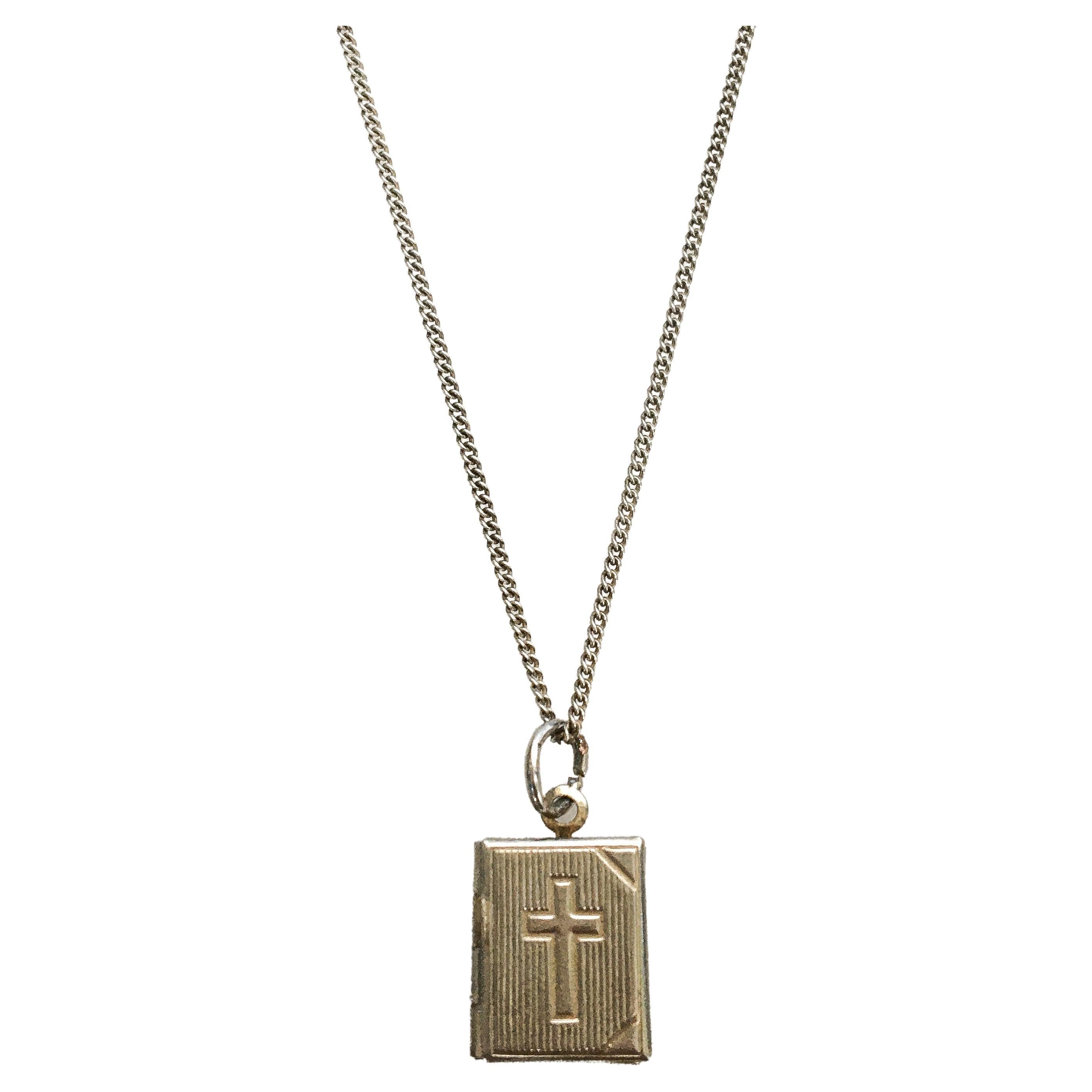 This lovely locket charm pendant is crafted in plated silver. The charm showcases a miniature book with a foxed and faded photo of mother Mary inside. On the front a cross is engraved while the locket itself has vertical engraved lines. The charm