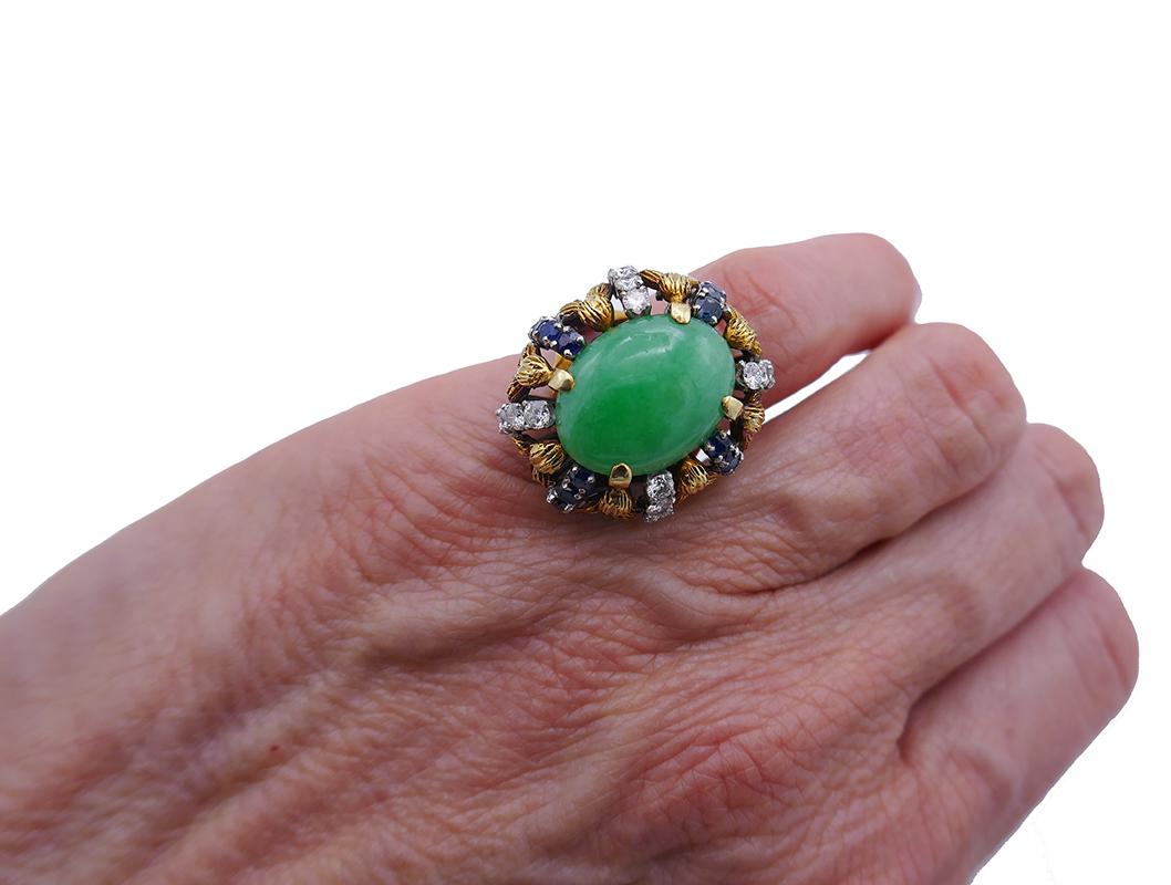         A head-turner cocktail ring made of 14k gold with jade cabochon, diamond and sapphire. 
	This gorgeous ring is a classic example of the 1960s jewelry. It’s big, well articulated and colorful. 
	The cabochon jade is skirted all around by the