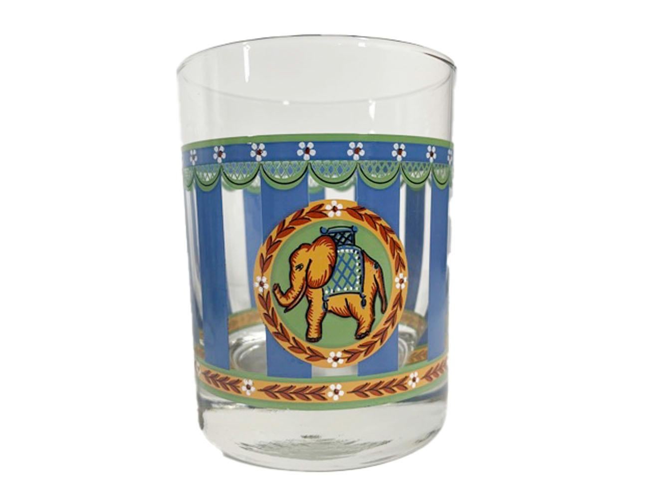 Vintage rocks glasses decorated with vertical blue bars with green tan and white decorative bands at top and bottom and having a medallion to one side with a caparisoned elephant within a wreath of leaves and flowers.