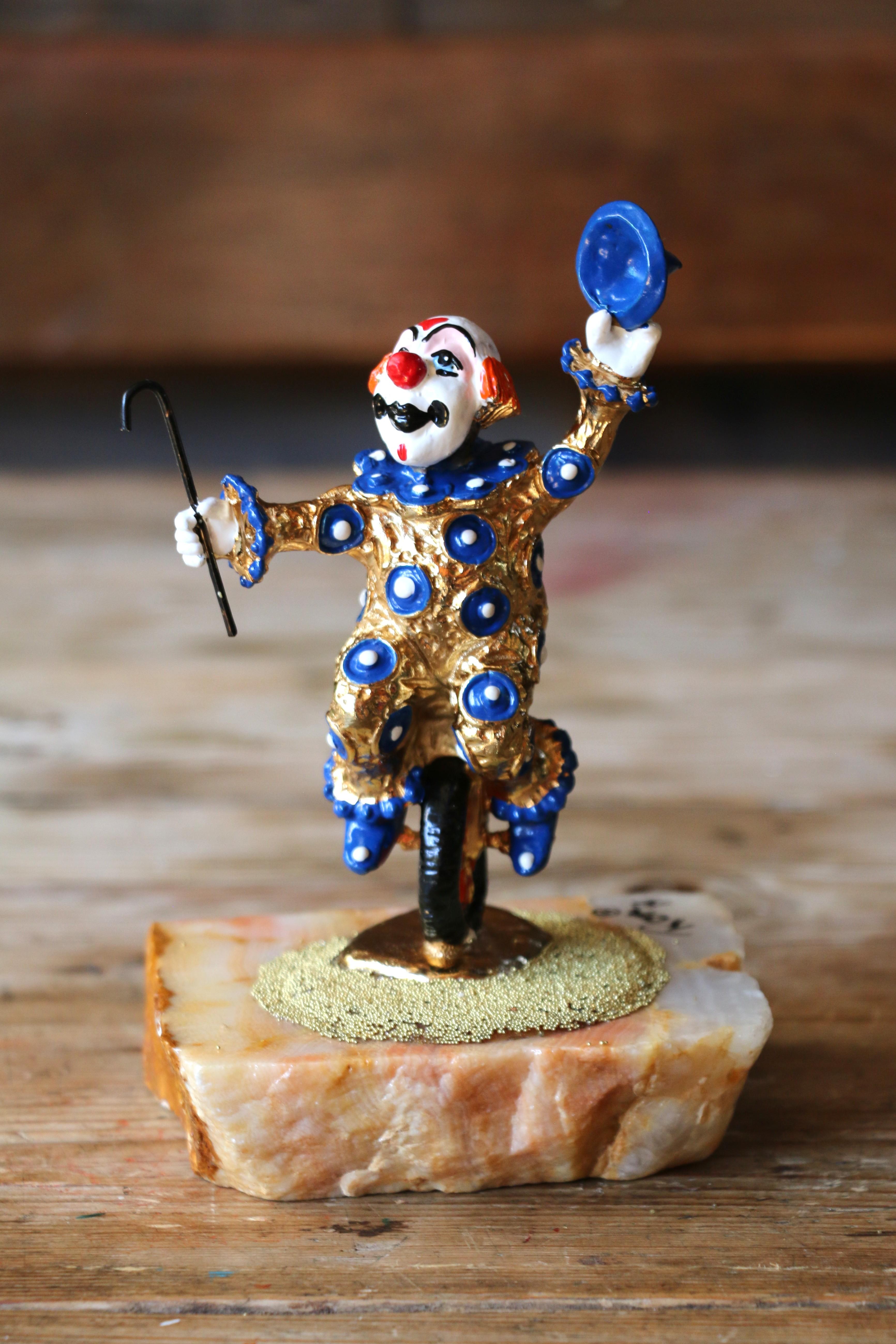 Splendida statuetta vintage creata da Ron Lee nel 1979. La statuetta raffigura un clown con un costume placcato oro a pois blu, che cavalca un monociclo e tiene in mano un bastone e un cappello blu, il tutto montato su una base in pietra lucida.