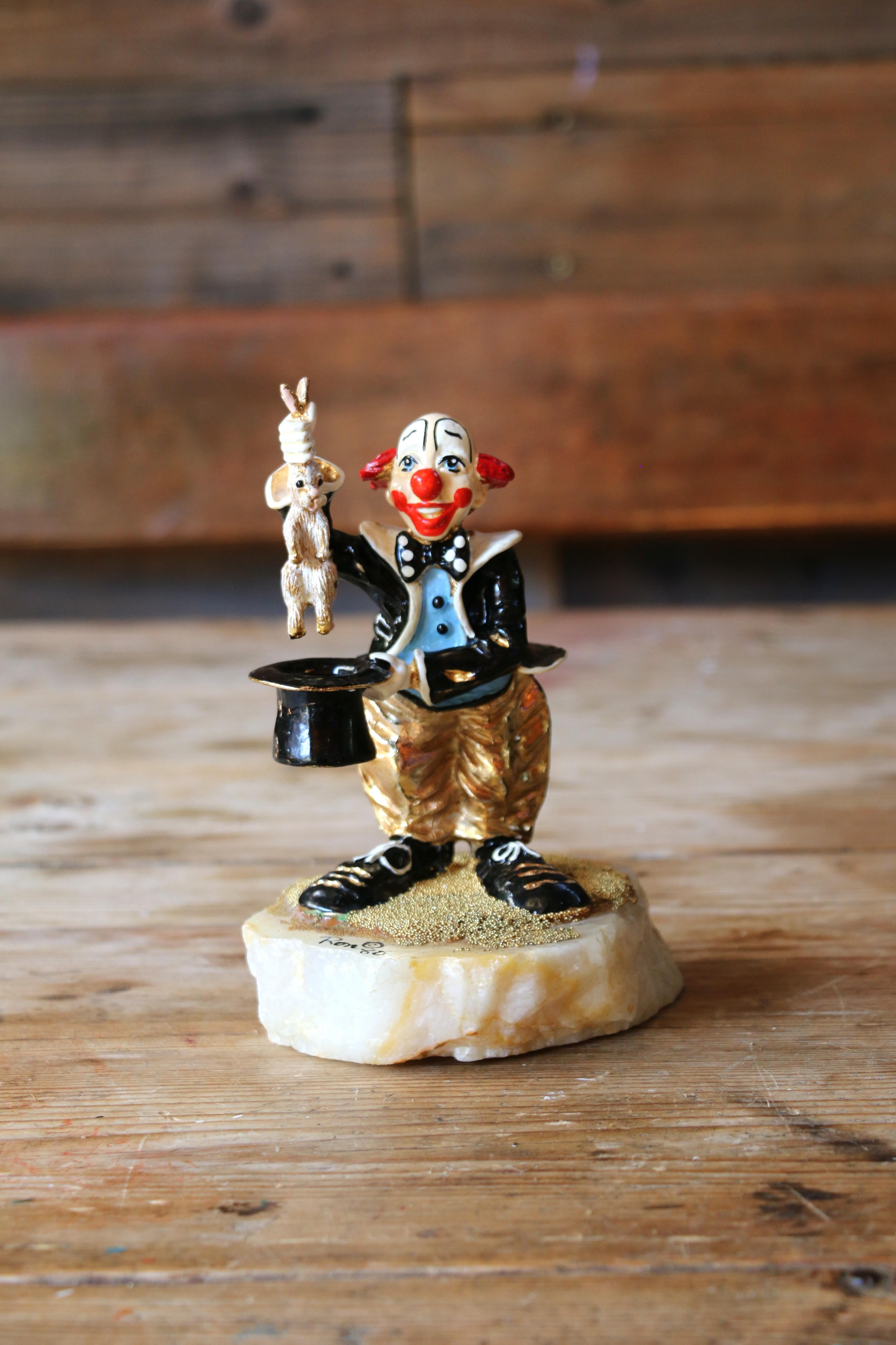 Splendida scultura vintage che raffigura un clown, vestito in nero e blu con jeans dorati, che esegue un trucco di magia estraendo un coniglio da un cappello a cilindro, il tutto su una base strutturata. Questo pezzo è firmato dall'artista e datato