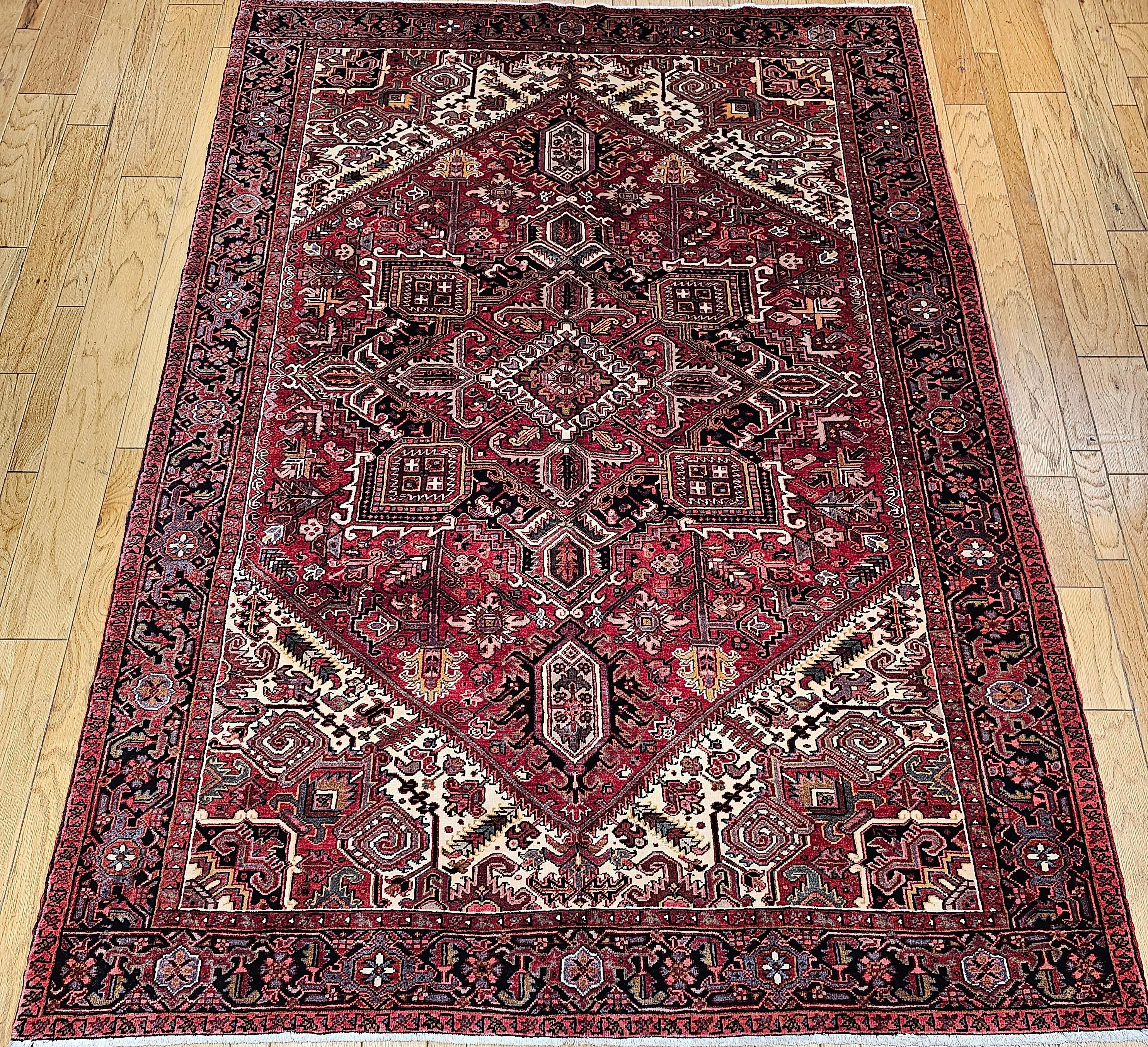 A room-size Persian Heriz rug from the 3rd quarter of the 1900s.  This Heriz has a brick-red field with a central medallion in dark blue and red.  The corner spandrels are in ivory color with geometric designs in yellow, green, blue, and red.  The
