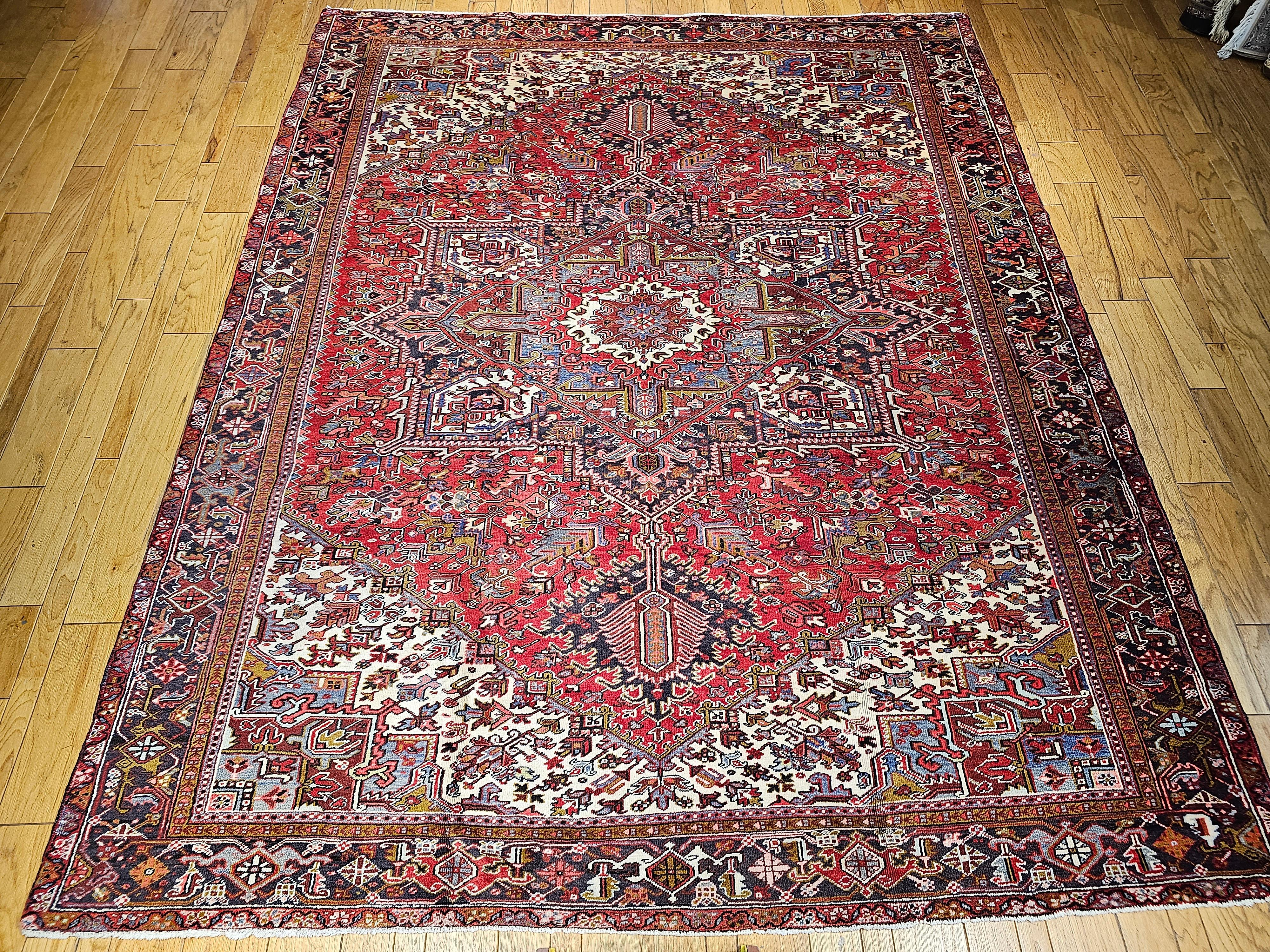 Room-size Persian Heriz in a medallion pattern in ivory, red, pale blue, yellow circa the mid 1900s.  The Heriz rug has a red field with a dark blue or lavender and ivory color spandrels.  The border is in a navy blue color.  The weave is very tight