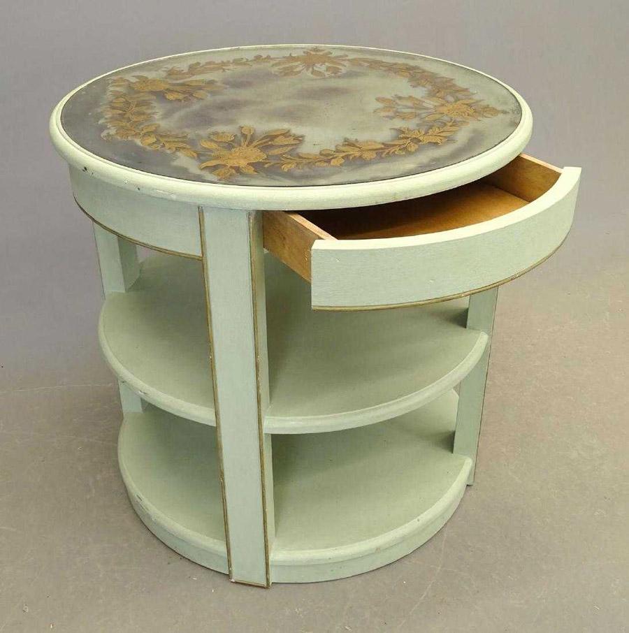 The vintage round side or end lamp table has a mirrored inset top embellished with a painted wreath of gold flowers and leaves. One section has a hidden drawer above the two shelves. Painted pale green with gilt trim. Could work great in any decor