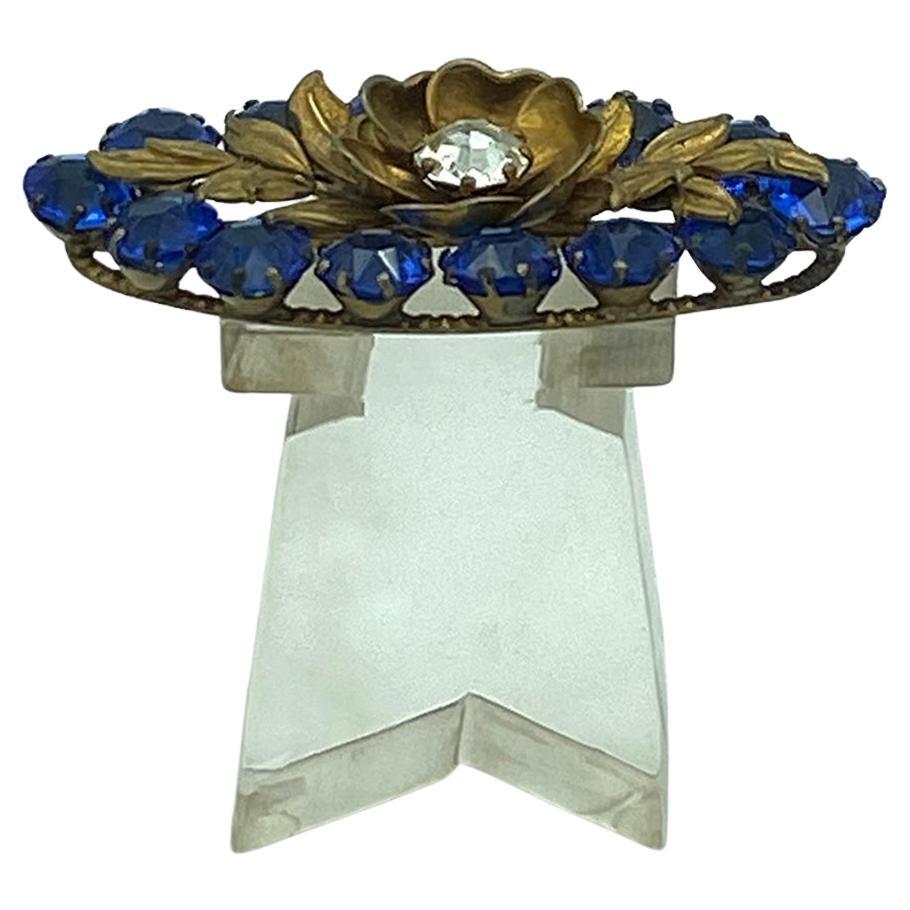 This is a 1930-40s royal blue crystal glass brooch. It has 14 pieces of 11mm faceted royal blue crystal stones surrounding a gilt flower with a 11mm clear rhinestone center. All stones are prong set on a structured bronzed ellipse shape ring.

Our