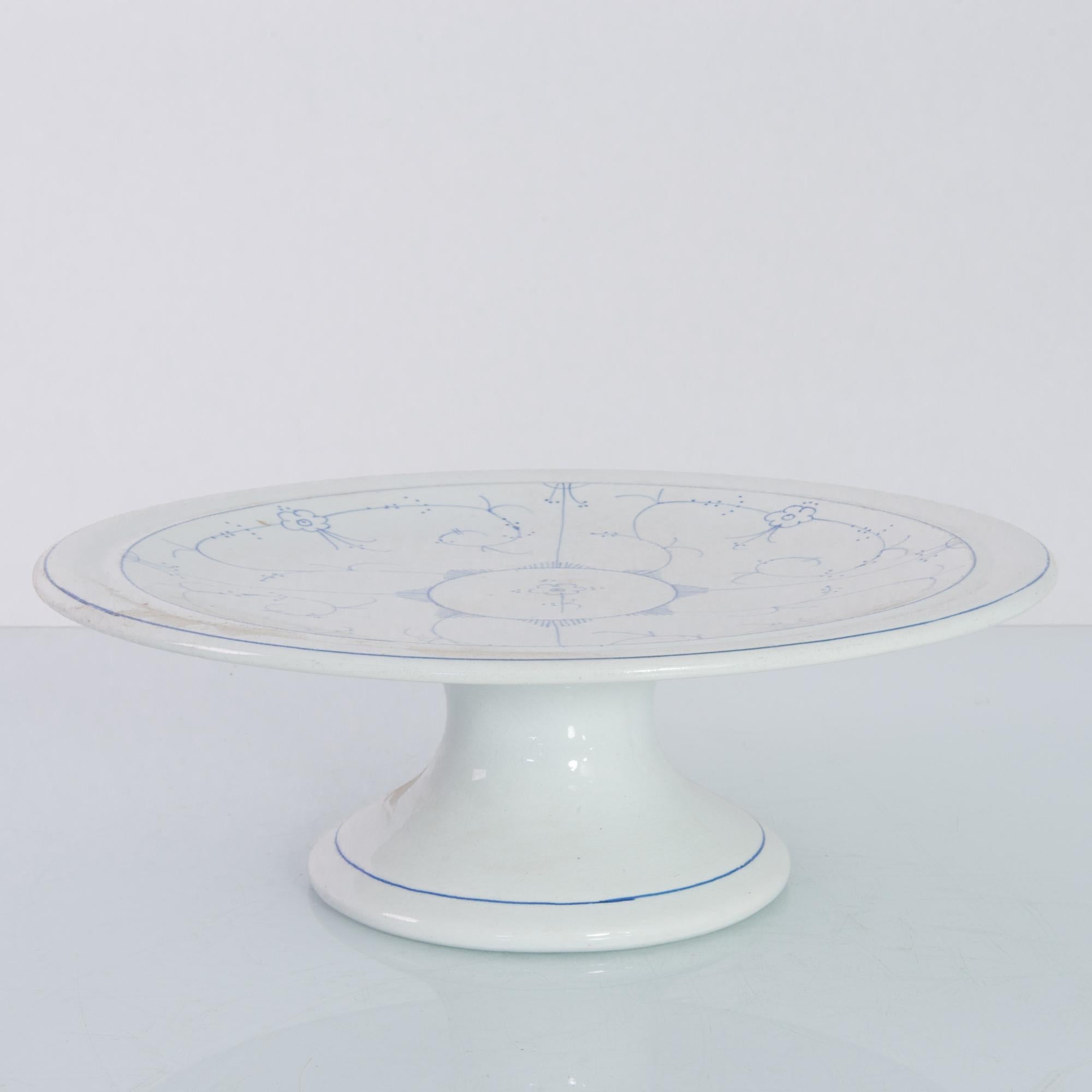 A ceramic cake tray from Denmark, produced circa 1900. A flat plane of circular ceramic coated in thick white glaze. Laced with the signature Royal Copenhagen blue painted glaze; this simple tray merely sits upon its stand and waits for cake. Only