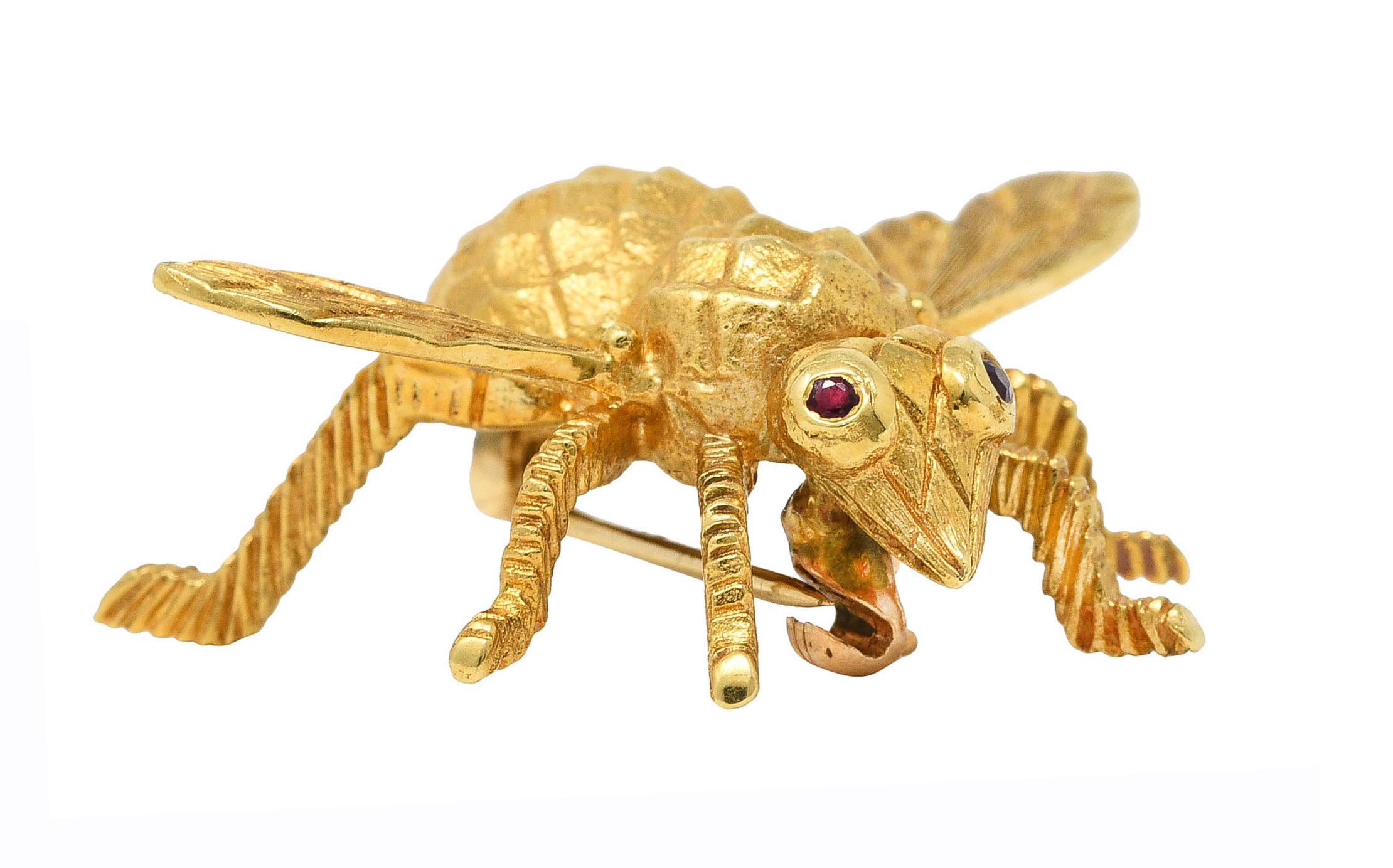 Brooch is designed as a stylized bug with textured body and engraved patterned wings

Accented by flush set round cut ruby eyes - transparent medium red in color

Completed by hinged pinstem with locking closure

Stamped 18k for 18 karat gold

With