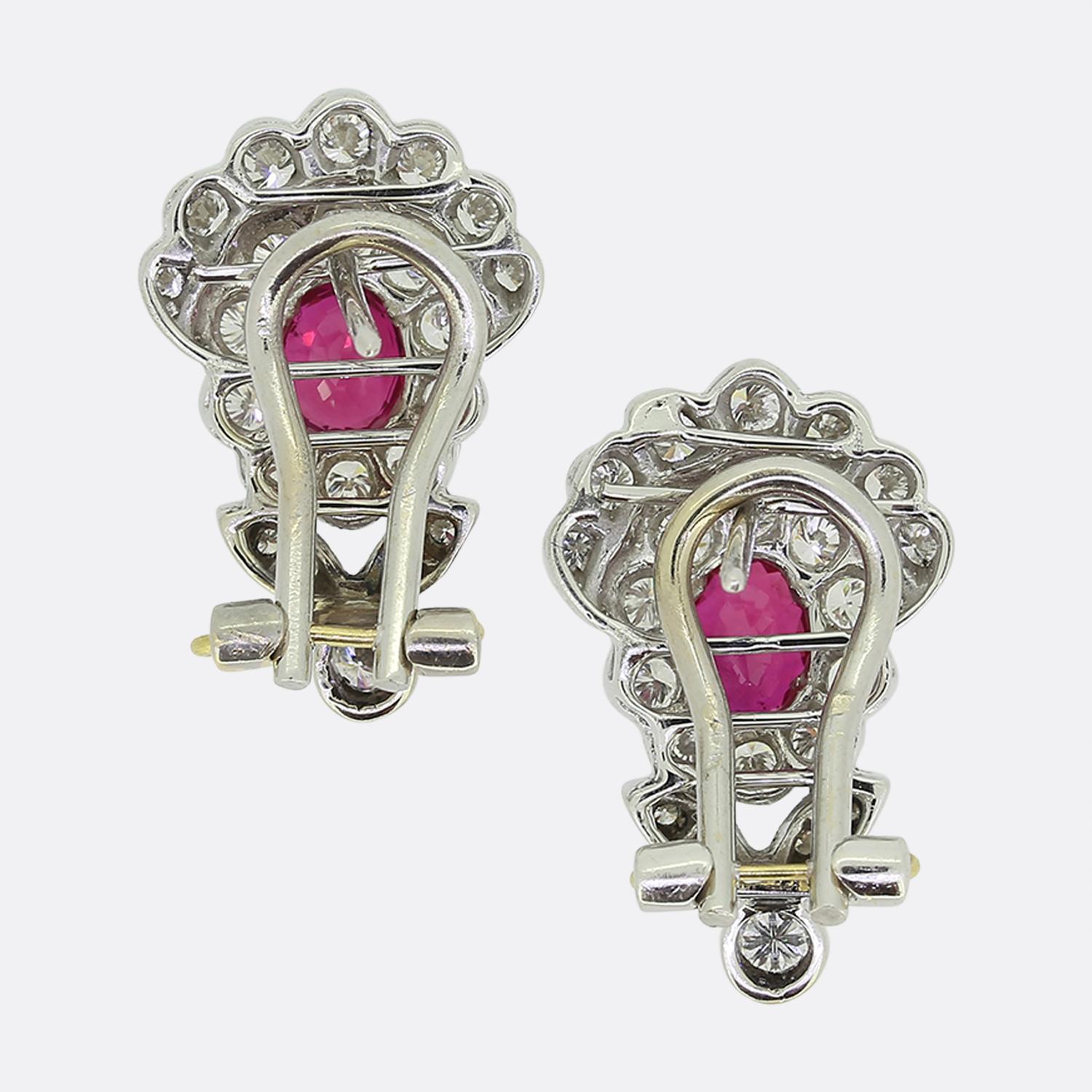 Here we have a charming pair of ruby and diamond cluster earrings. Each identical piece has been crafted from 18ct white gold and features a single oval faceted ruby possessing a rich vivid red colour tone. This principal stone sits slightly risen