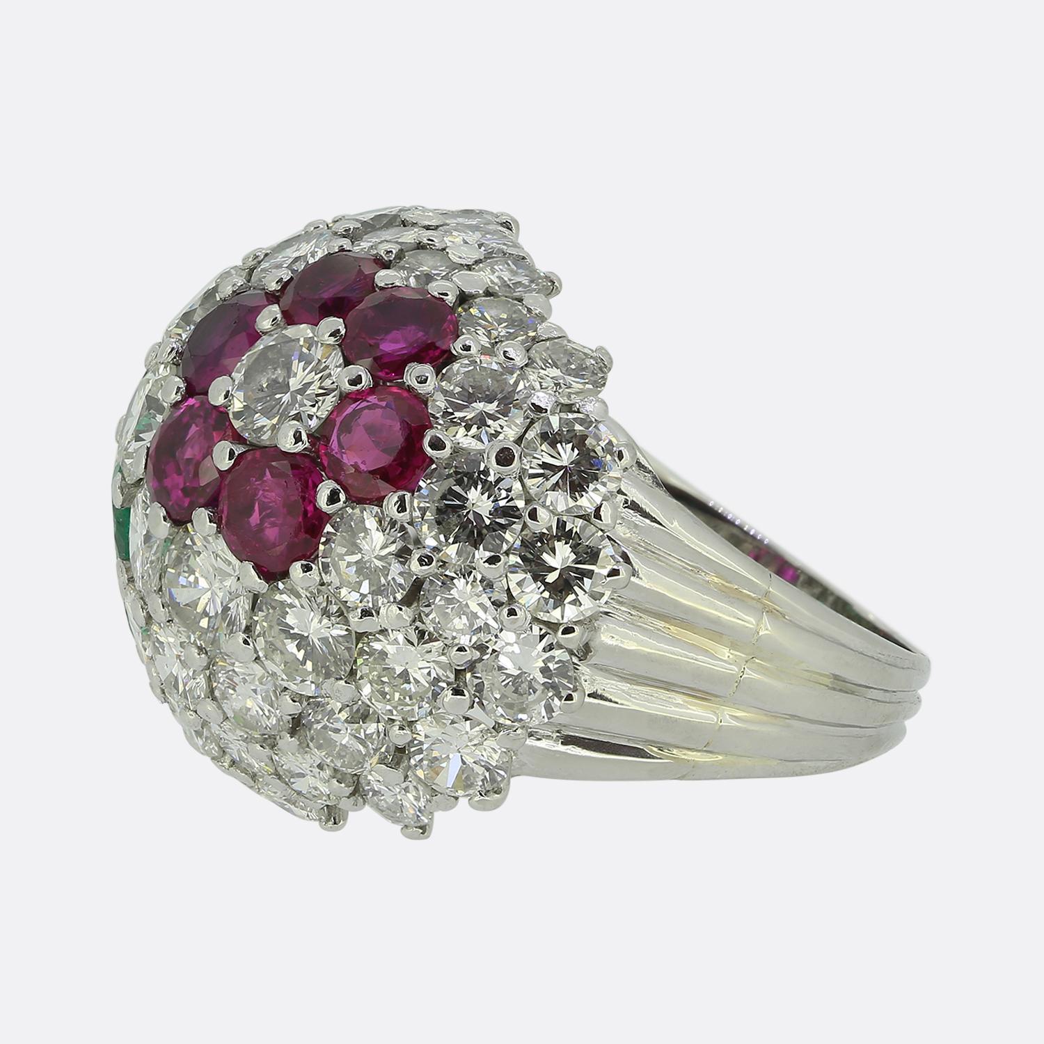 Here we have a stunning bombe ring featuring a vibrant array of gemstones. A domed shaped head plays host to clusters of perfectly matched round red rubies and green emeralds, interspersed with bright white brilliant cut diamonds. All stones have