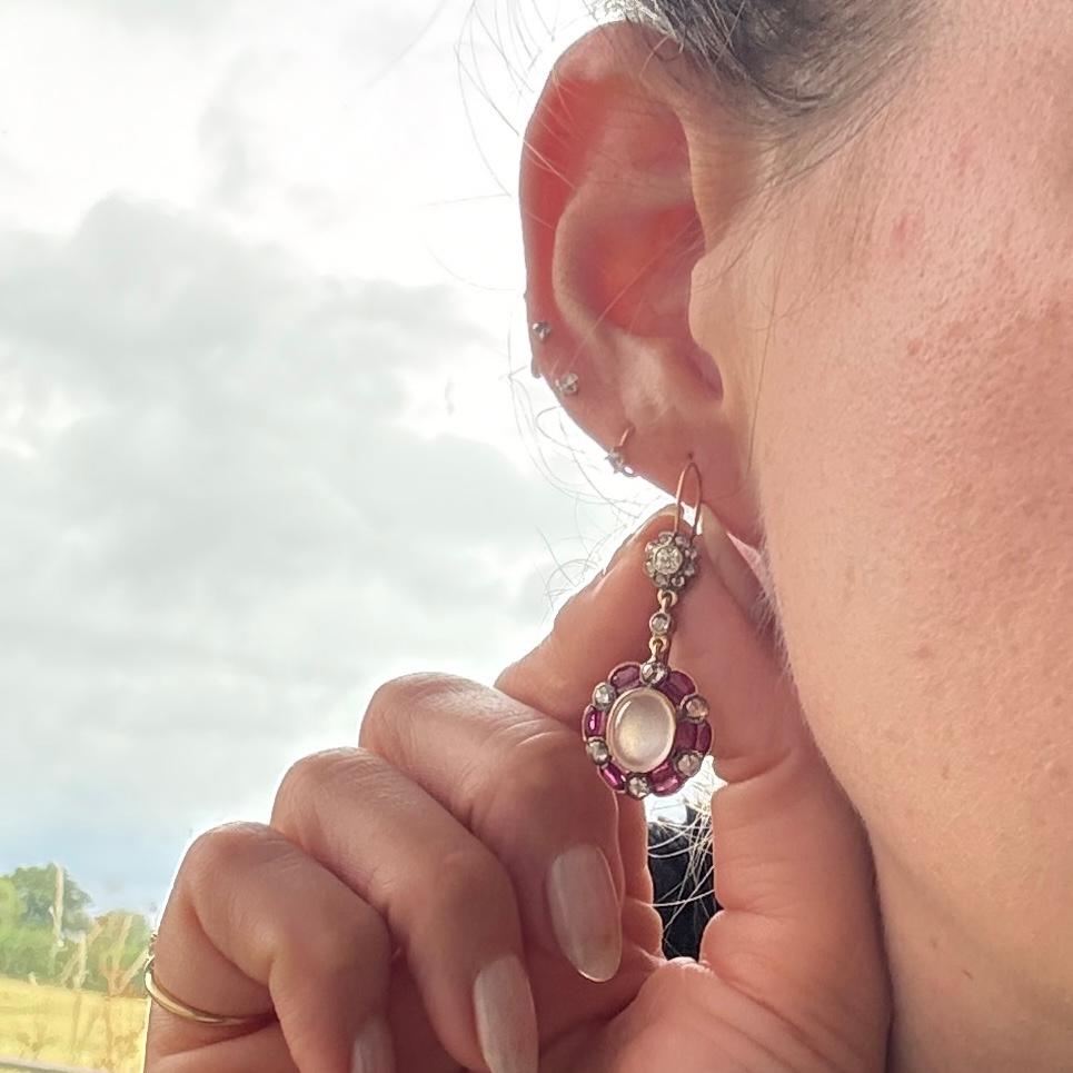 Cada pendiente contiene una piedra lunar con un hermoso brillo azul.  Alrededor de esta magnífica piedra hay un halo de rubíes y diamantes talla rosa. Los pendientes están modelados en metal amarillo. El total de rubíes de cada pendiente es de 60